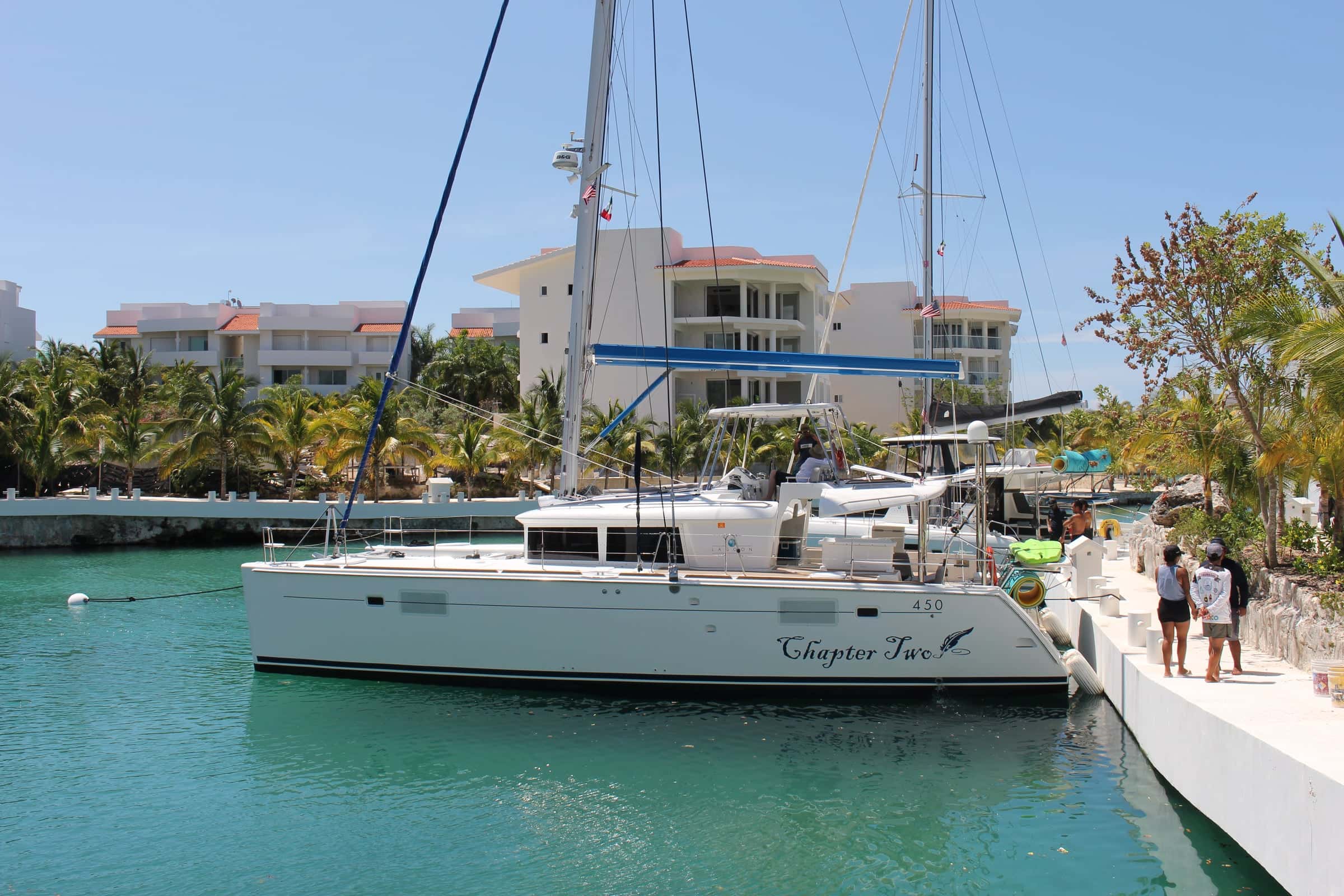 The catamaran moored along the marina quay, her name on the hull, palms and white apartments behind