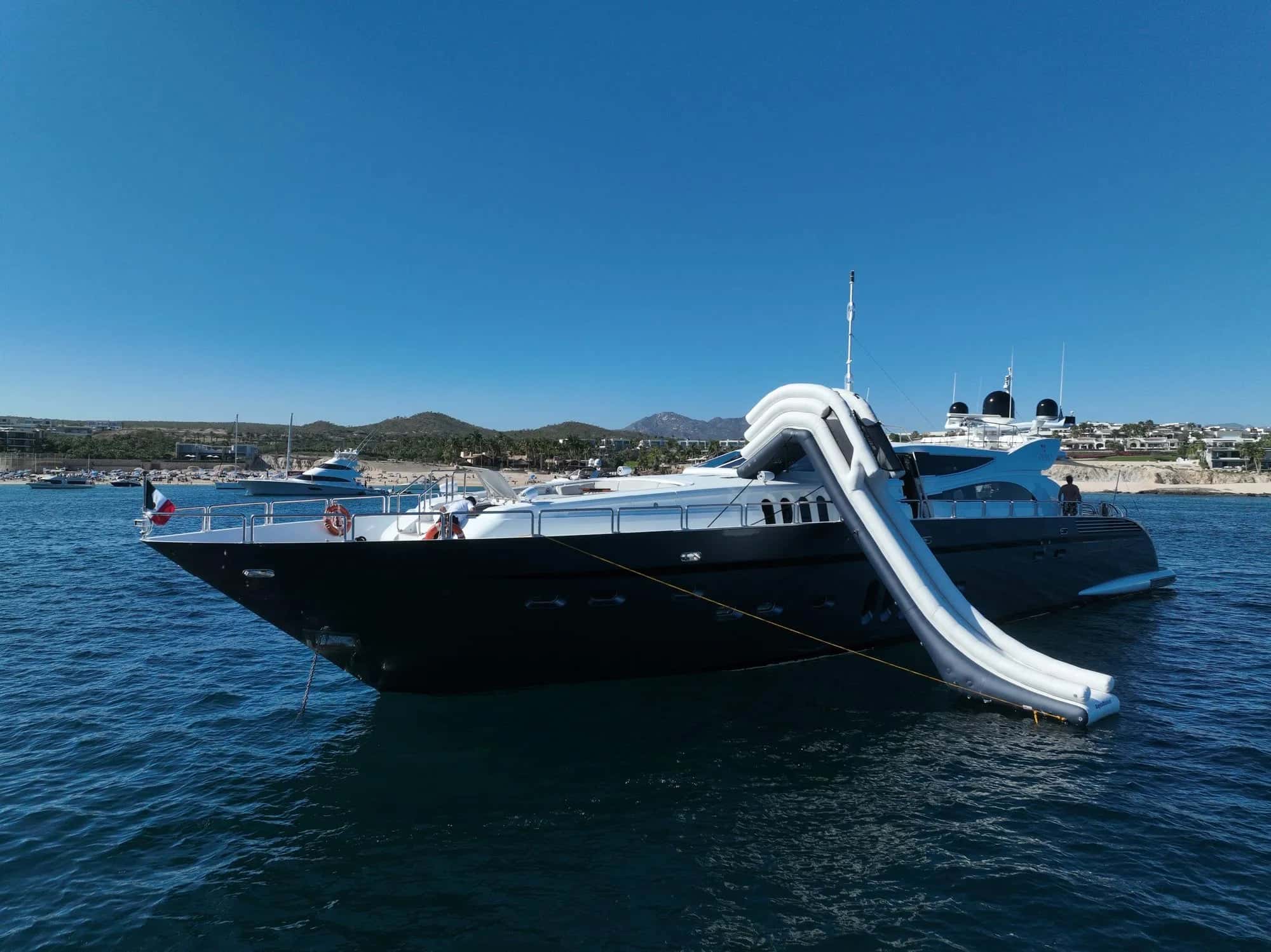The yacht at anchor with an inflatable slide running from the upper deck into the sea, hills and a marina behind