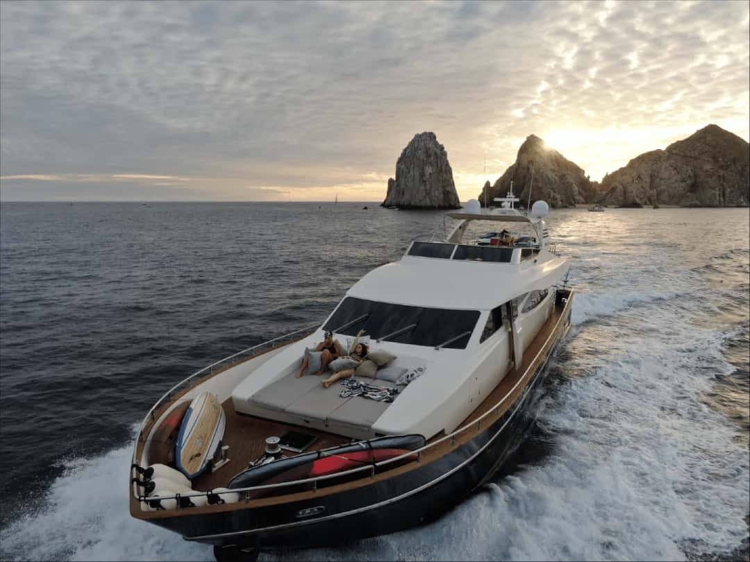 Black-hulled motor yacht seen from the front and above at sunset, two people lounging on the bow sunpad
