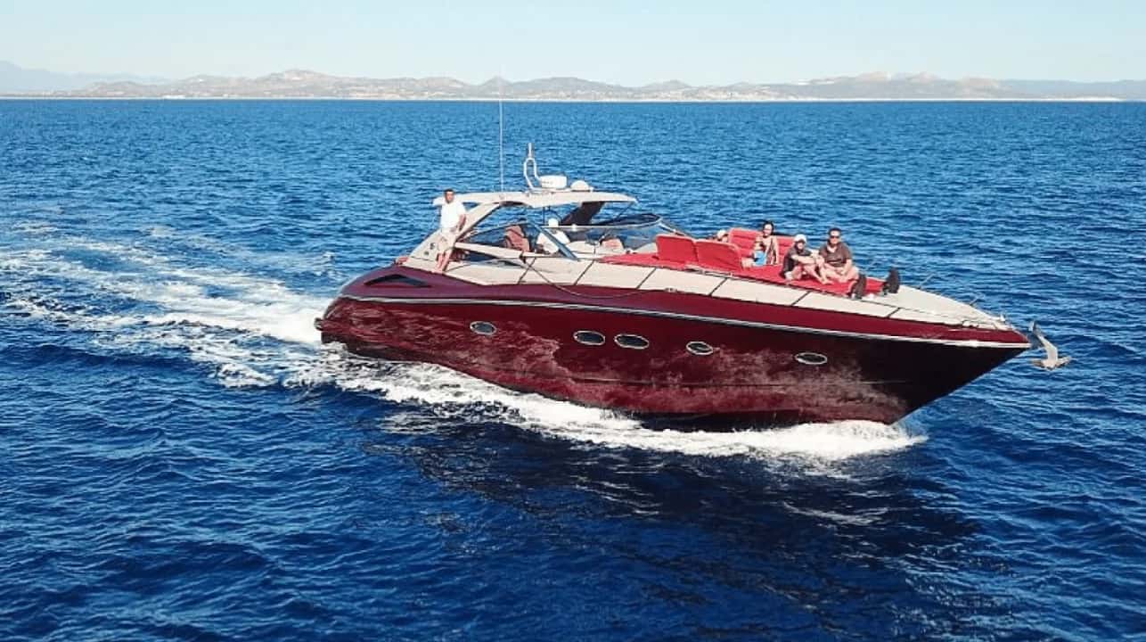 Dark red motor yacht cruising at speed on deep blue water, with guests seated on the red bow cushions