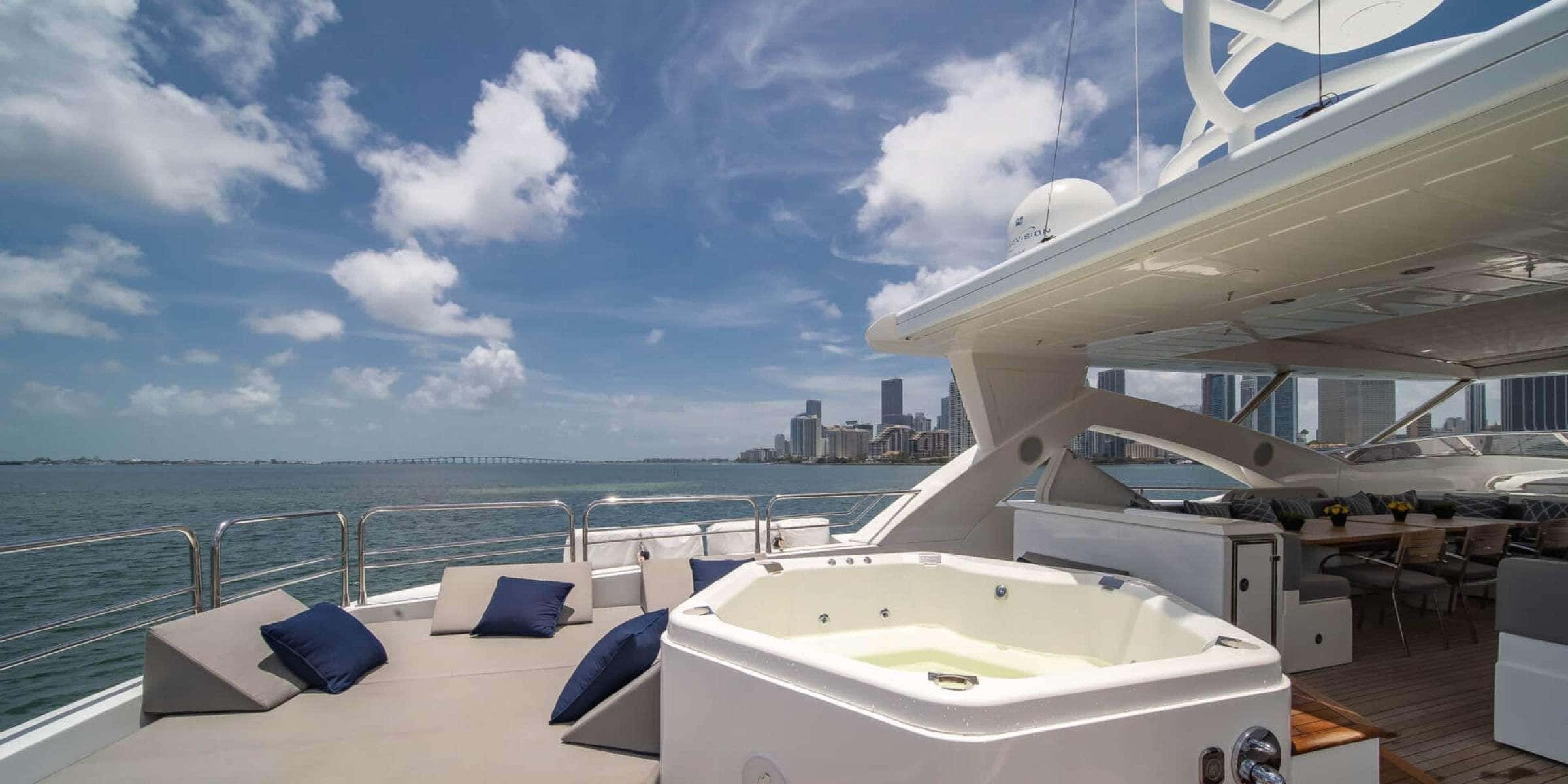 Sun pad with cushions and a hot tub on the aft deck, with a covered dining table and the Miami skyline behind
