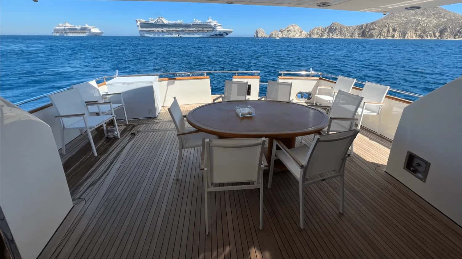 Aft deck with a round teak table and white chairs, cruise ships and rocky coast beyond