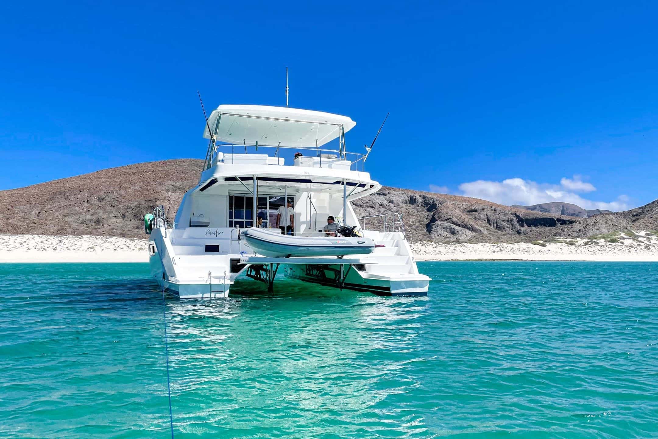Catamaran seen from the stern with a tender on the davit platform in turquoise water off a white beach