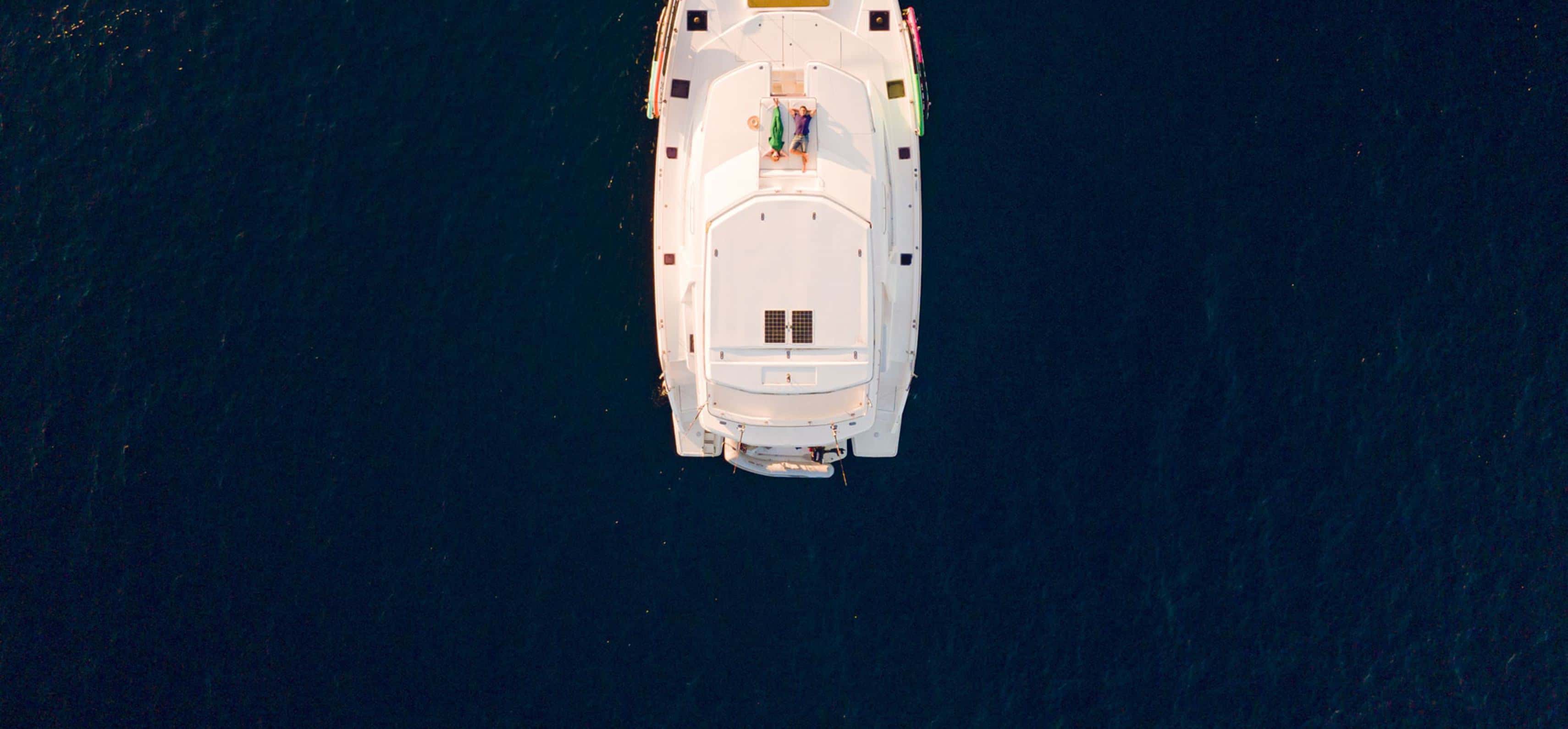 Overhead aerial view of a catamaran from the bow in dark blue water, two people sunbathing on deck