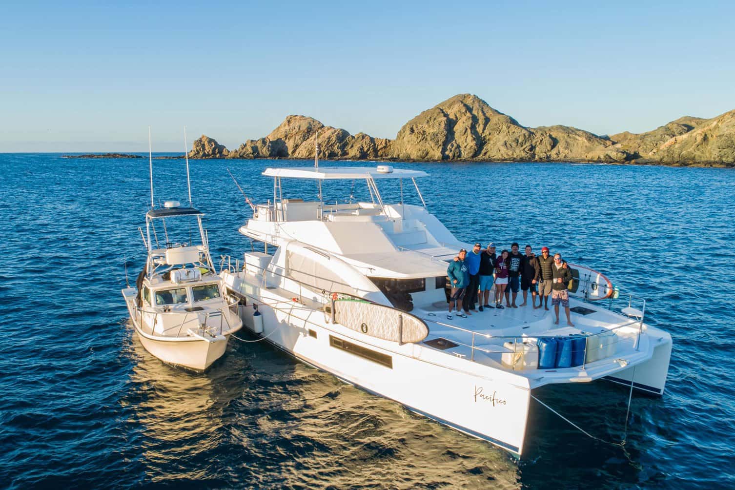 Catamaran Pacifico at anchor with a small fishing boat alongside and eight people standing on the aft deck
