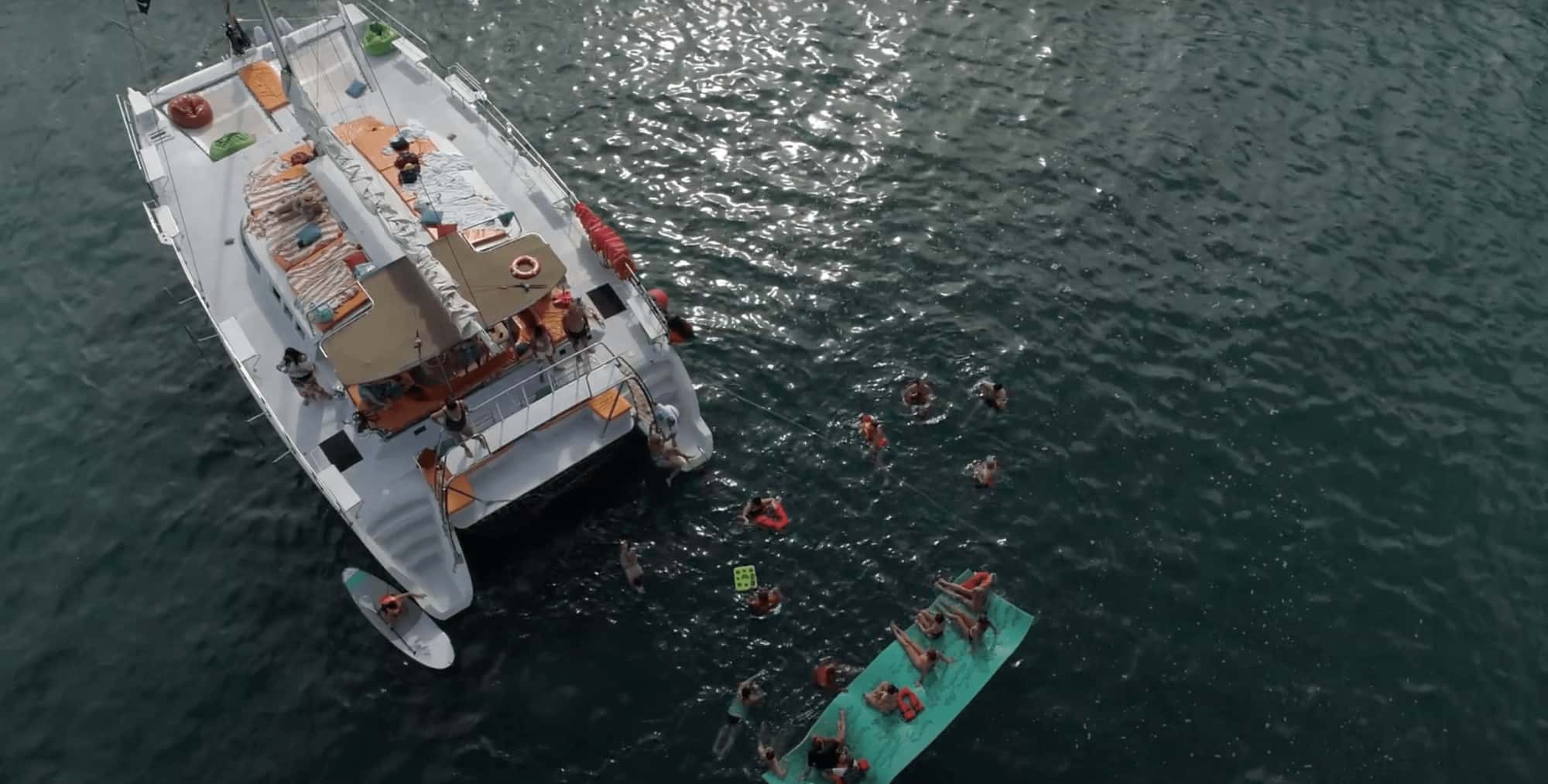 Overhead view of the catamaran with sun pads, bean bags and swimmers on a floating mat beside it