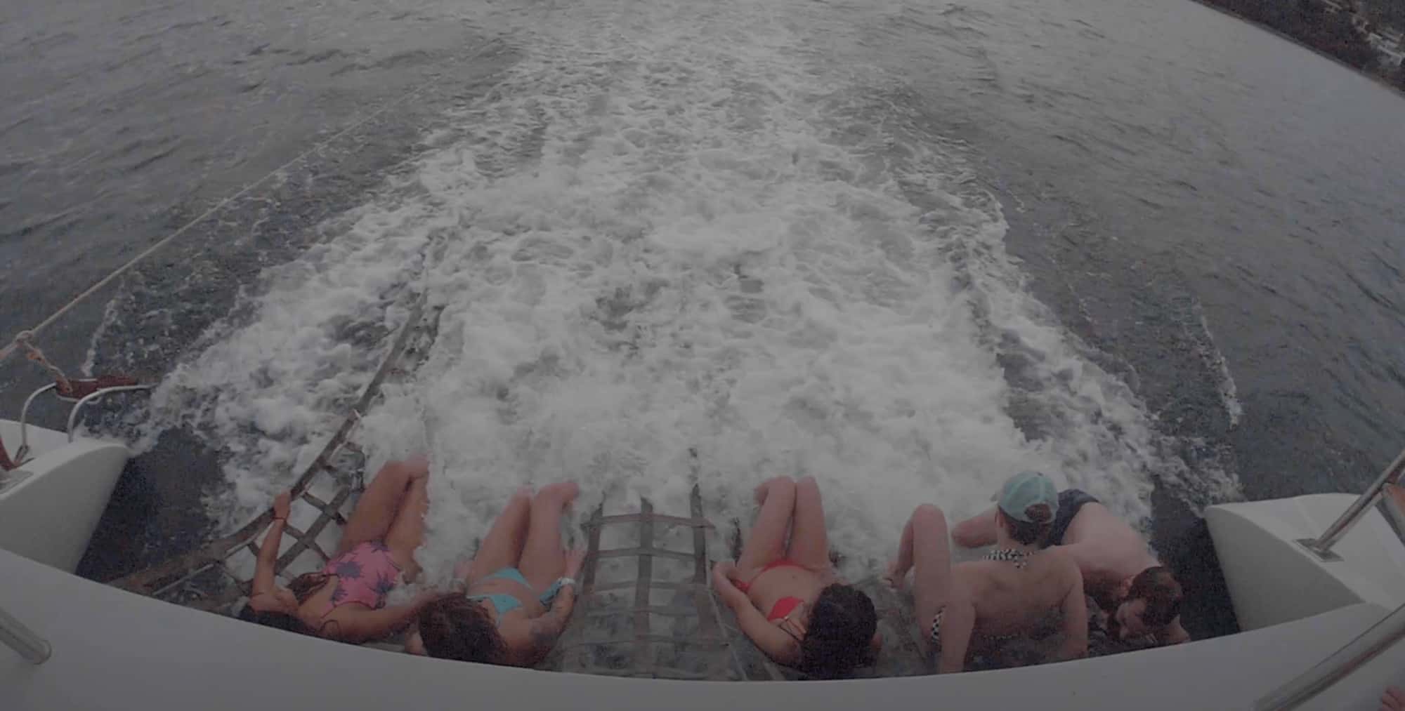 Guests sitting on the rope net at the back of the catamaran above the churning wake