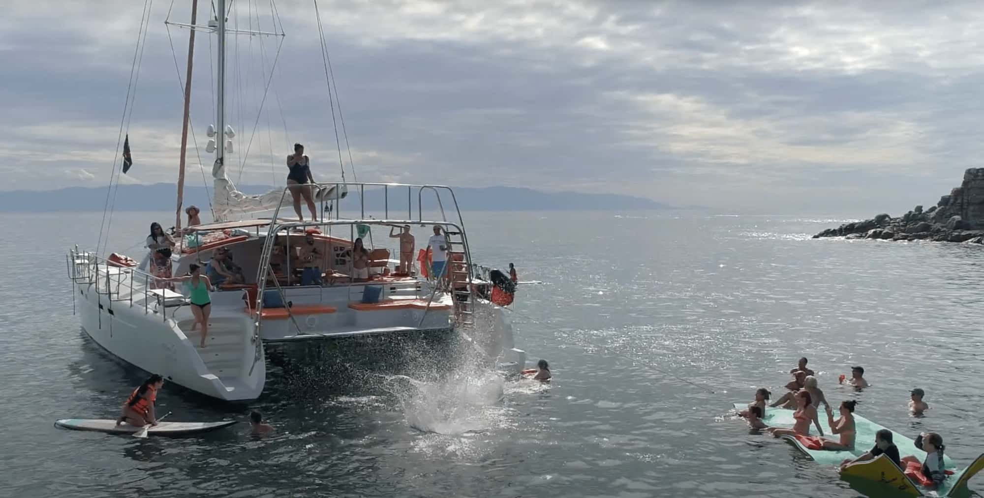 Ally Cat catamaran from the stern, a guest splashing into the sea, paddleboard and floating mat nearby