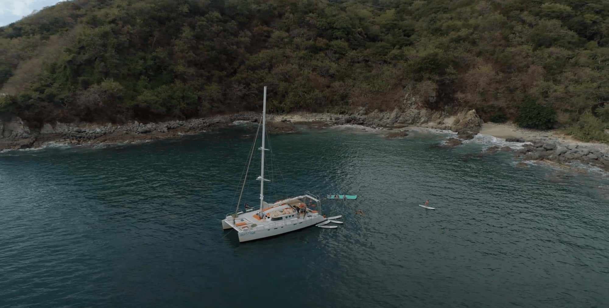 White catamaran at anchor in a green cove, with paddleboards and a floating mat in the water