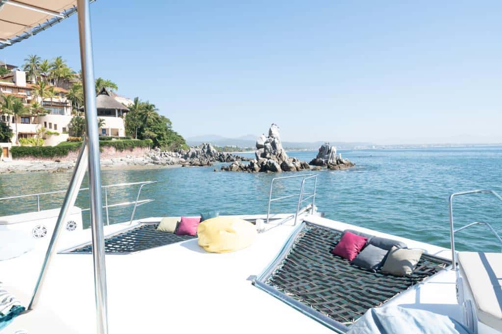 Bow deck with two netting areas, cushions and a bean bag, facing a rocky coast and villas