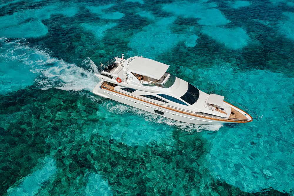 High aerial view of the white yacht under way over clear turquoise reef water, leaving a wake