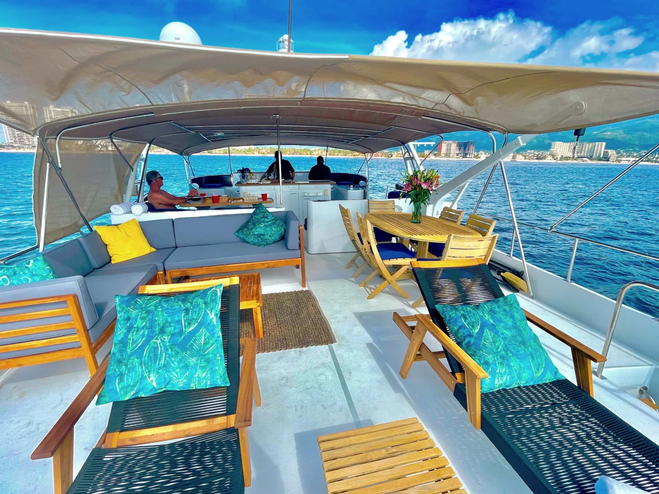 Upper deck with a grey sofa, loungers, an oval teak dining table and crew at the galley