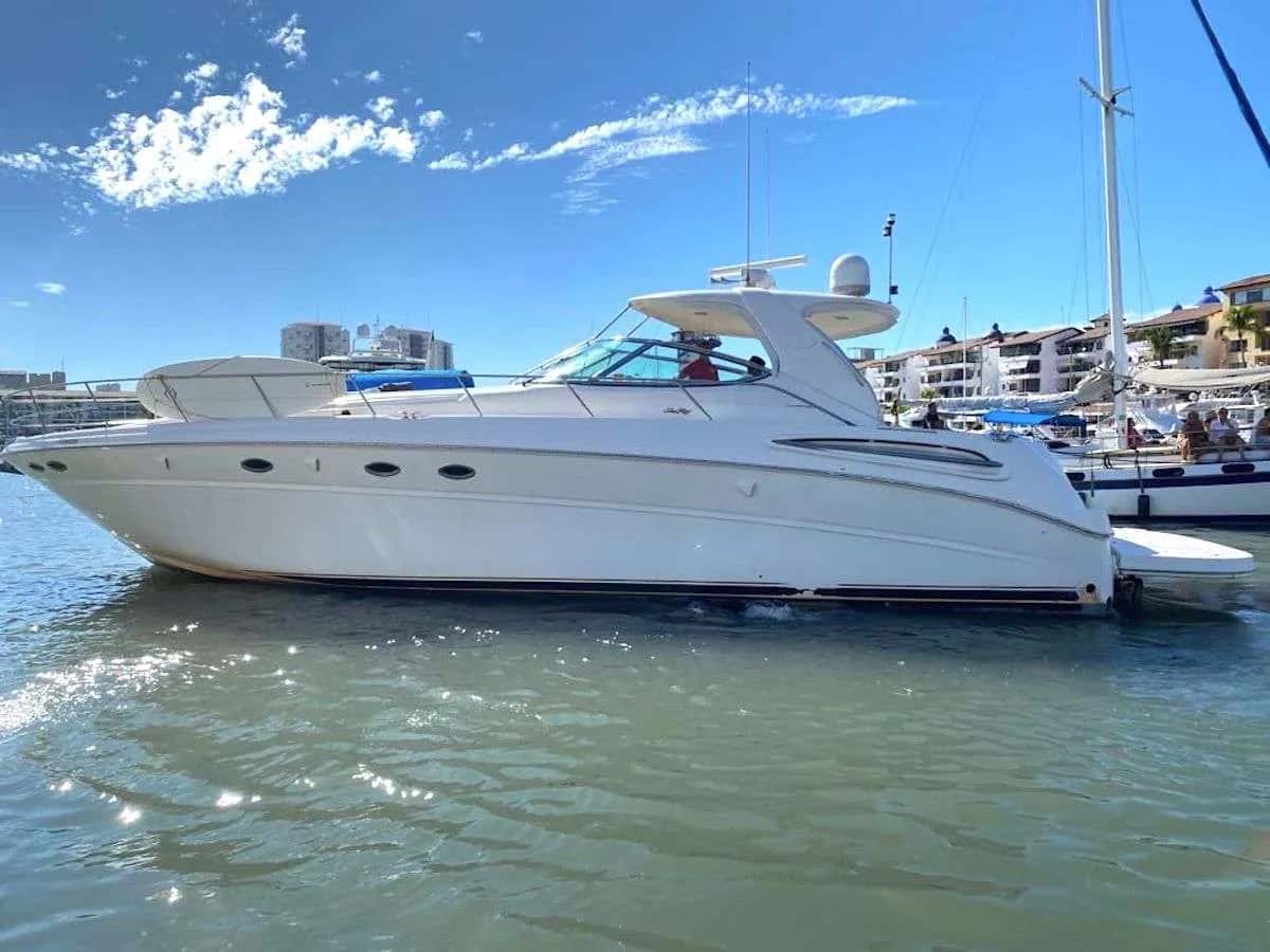 White motor yacht seen from the side in a marina, with blue sky and moored boats behind