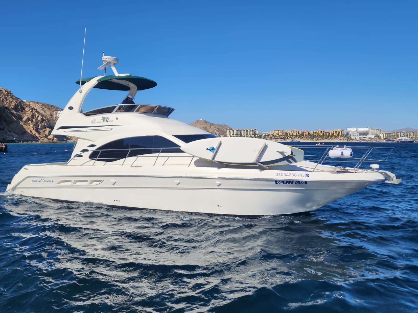 Vaduna, a white flybridge motor yacht under way in blue water off Cabo, a paddle board on deck