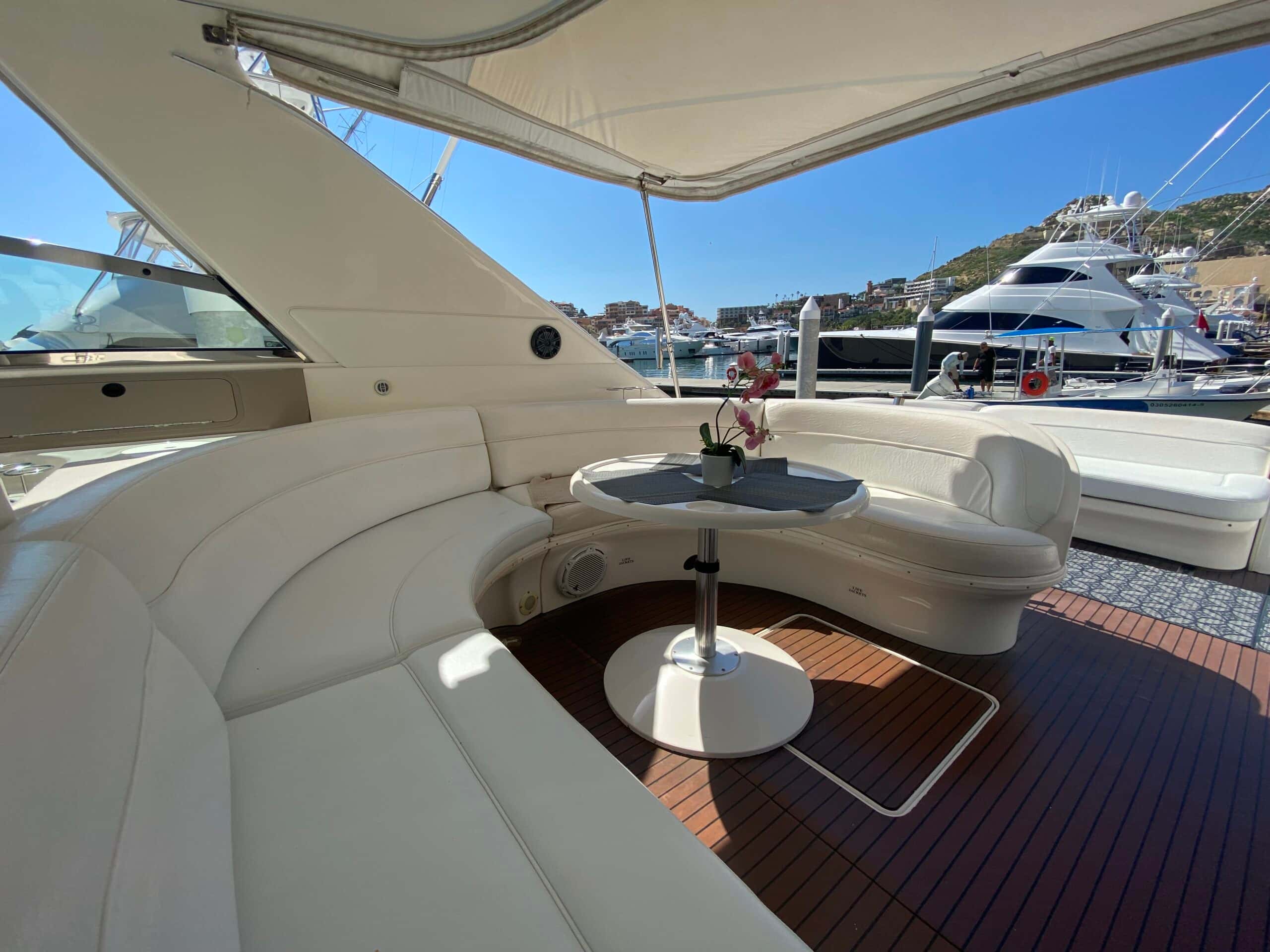 Curved white seating around a pedestal table under a bimini on a teak cockpit, with a marina and yachts behind