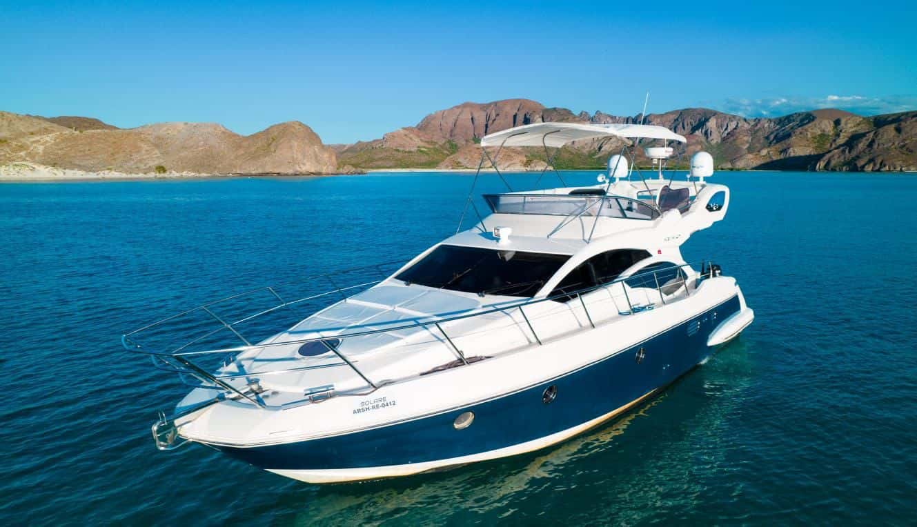 Golden at anchor in blue water off a rocky shore, blue and white hull with flybridge and bimini