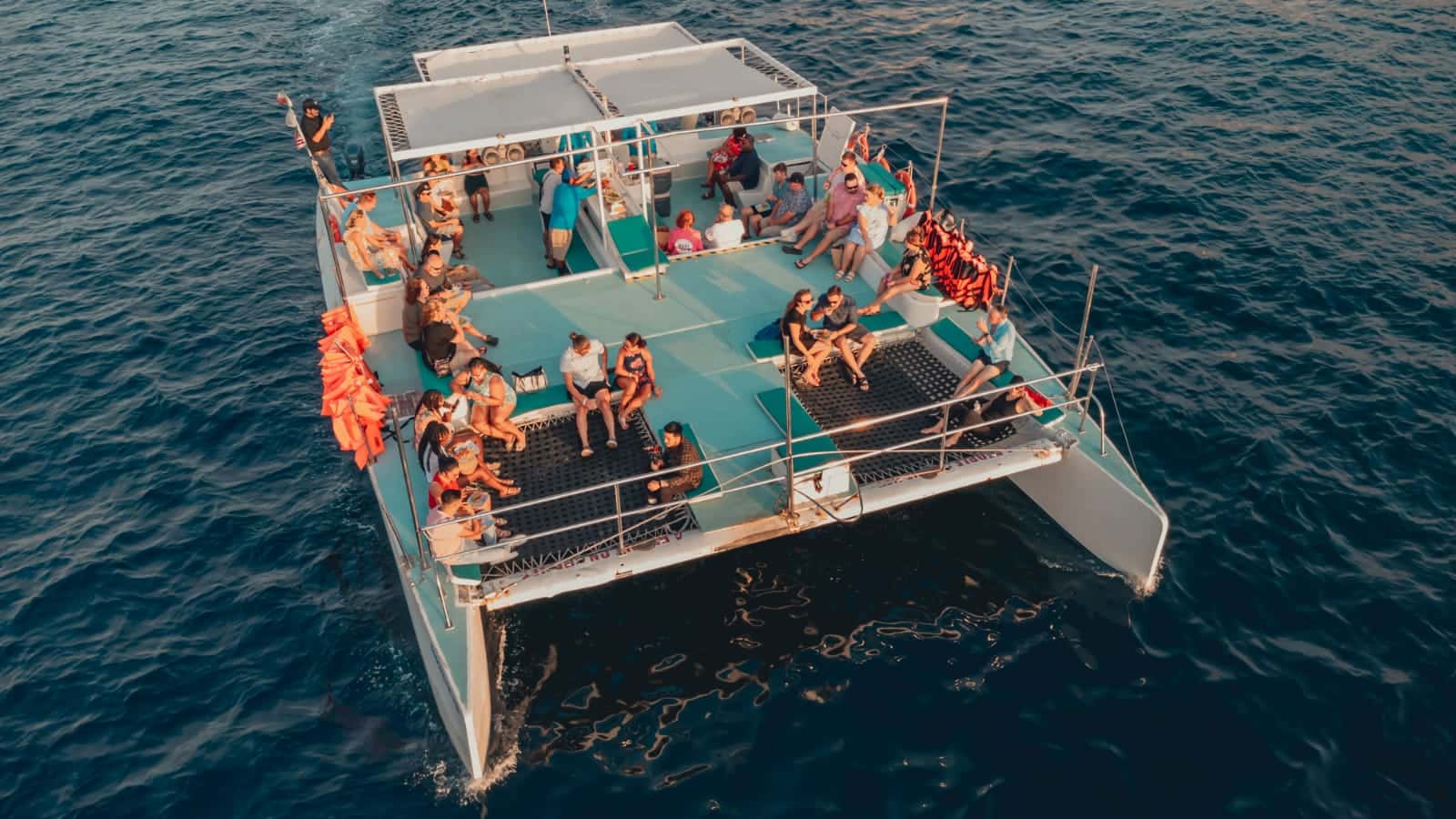 Aerial view of a catamaran deck, guests seated on cushions and trampoline nets, life jackets at the rail