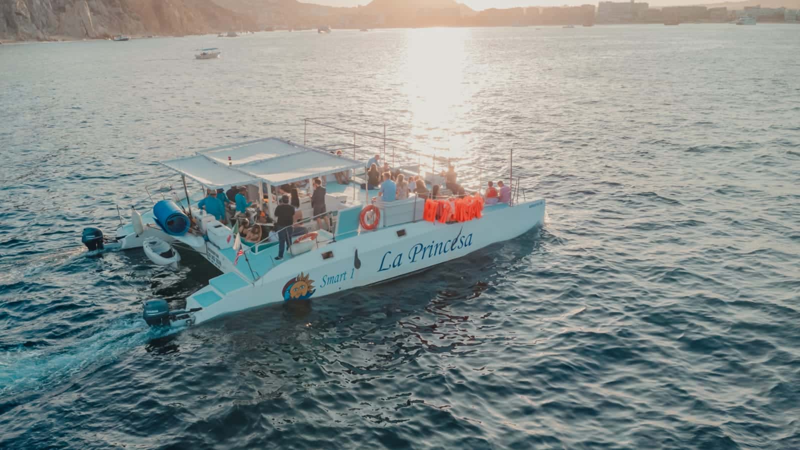 Catamaran with a shaded deck and guests aboard, seen from above at sunset off the Cabo coast