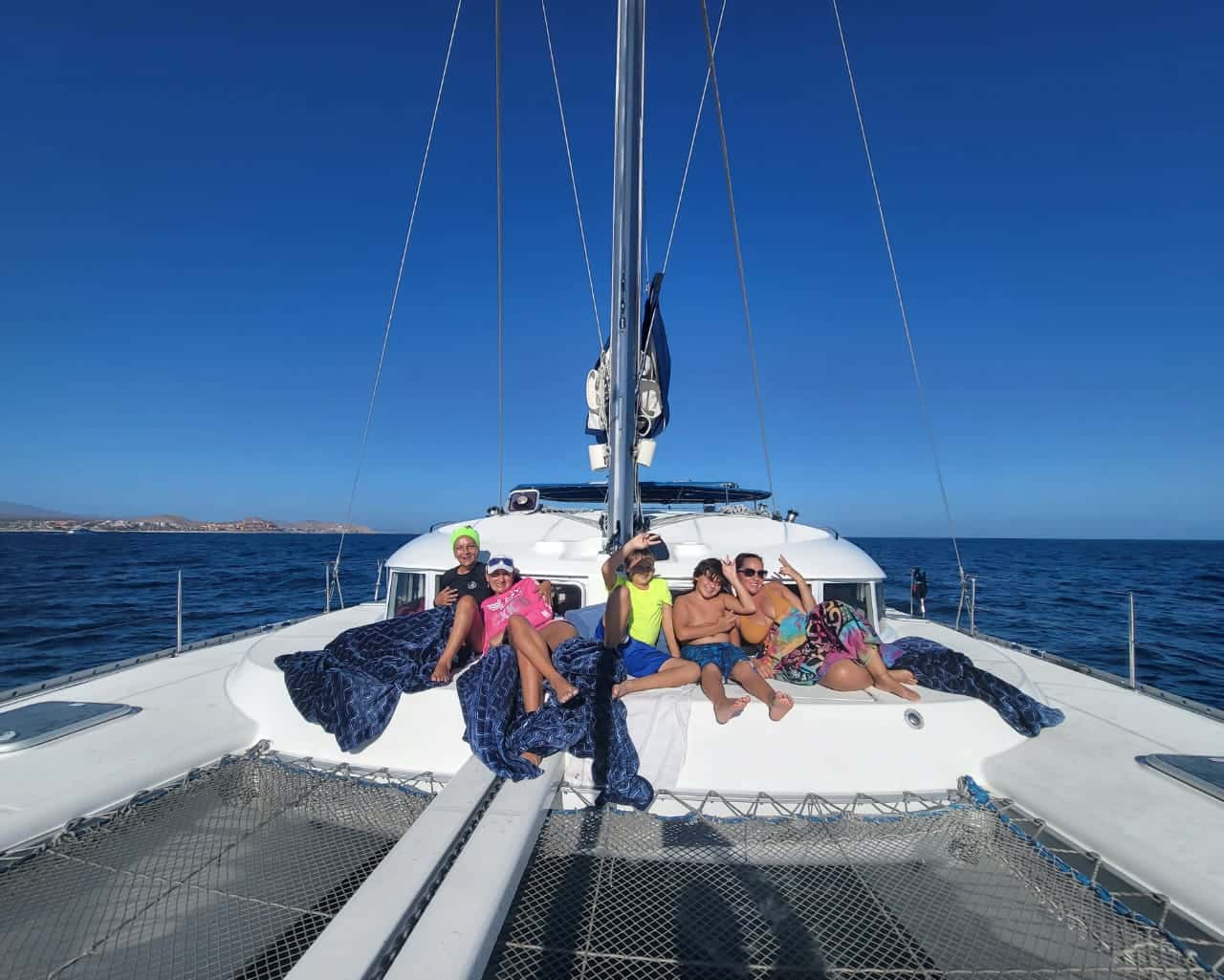 Guests resting on a blue blanket on the bow of a catamaran, nets below, coast in the distance