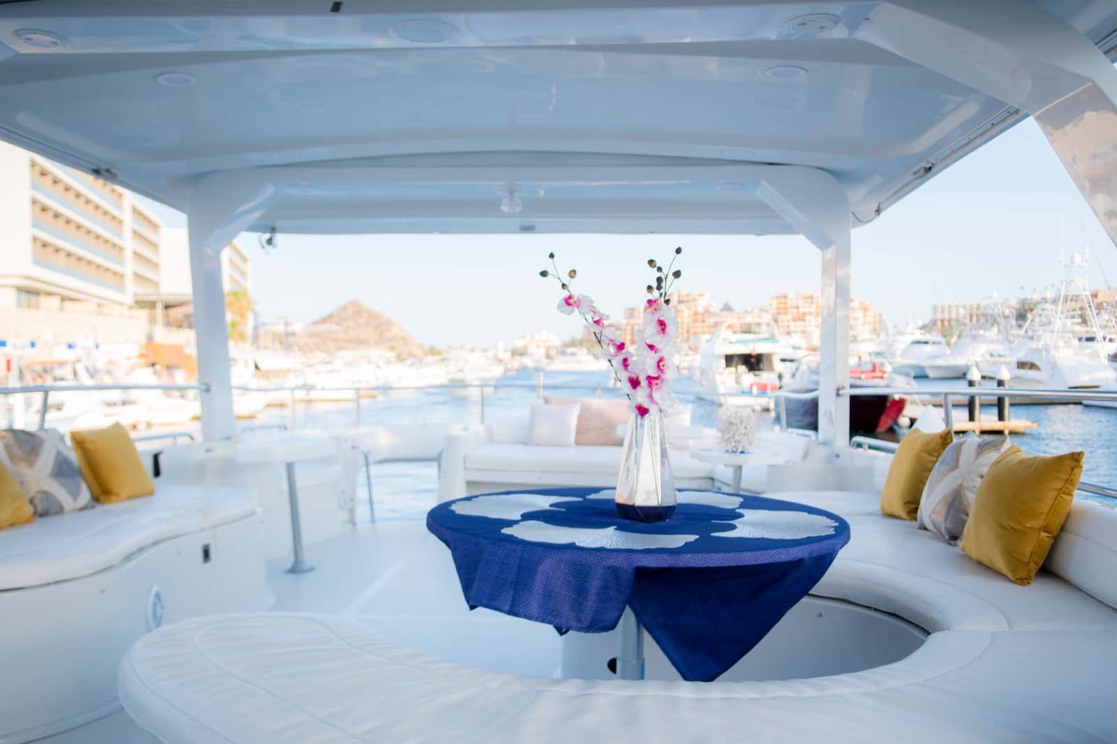 Shaded upper deck with white cushioned seating, yellow pillows and a round table with orchids, marina behind
