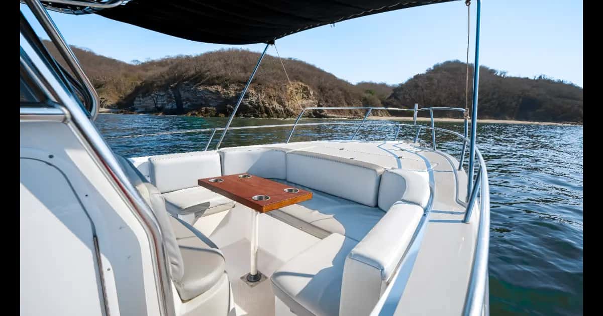Bow lounge with white cushioned seats and a teak cup-holder table, hills and beach behind