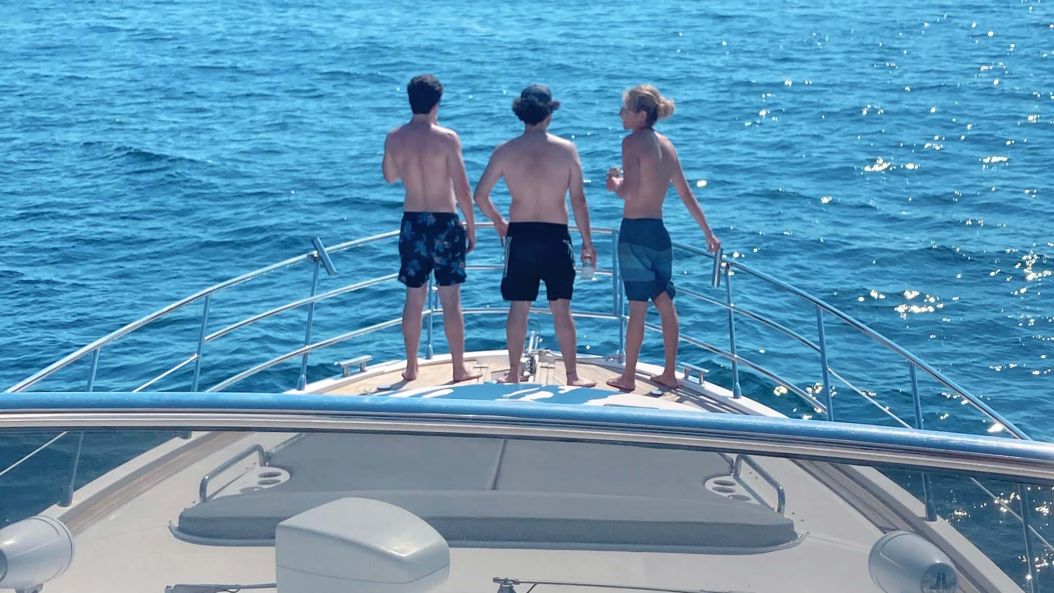 Three men in swim shorts standing at the bow rail looking out over open blue water