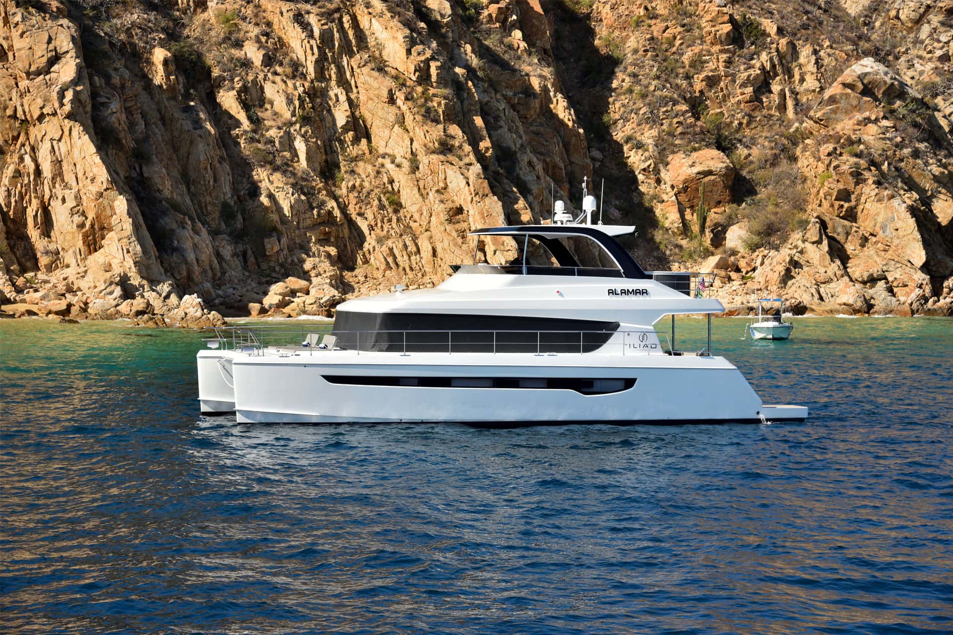 White Alamar catamaran at anchor below orange rock cliffs, a small boat beyond