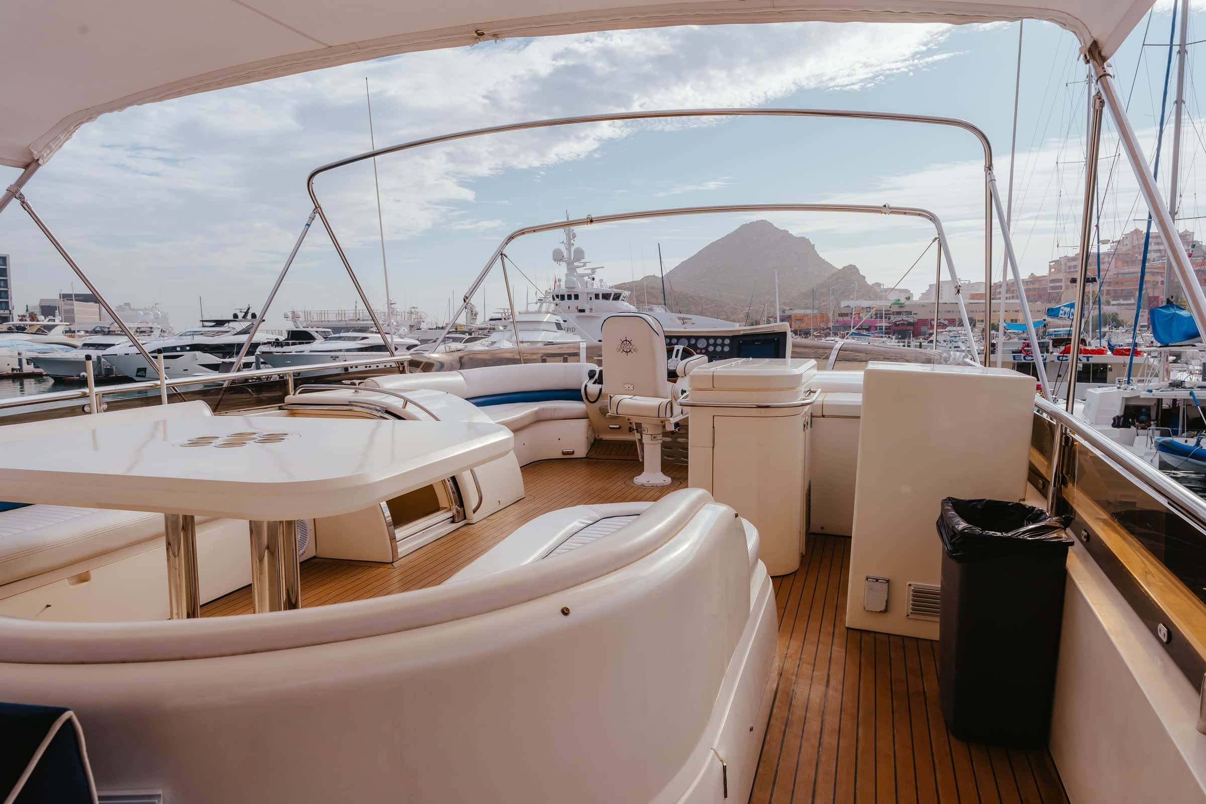 Flybridge under a canopy, the upper helm beside white curved seating and a table, the marina and a hill beyond