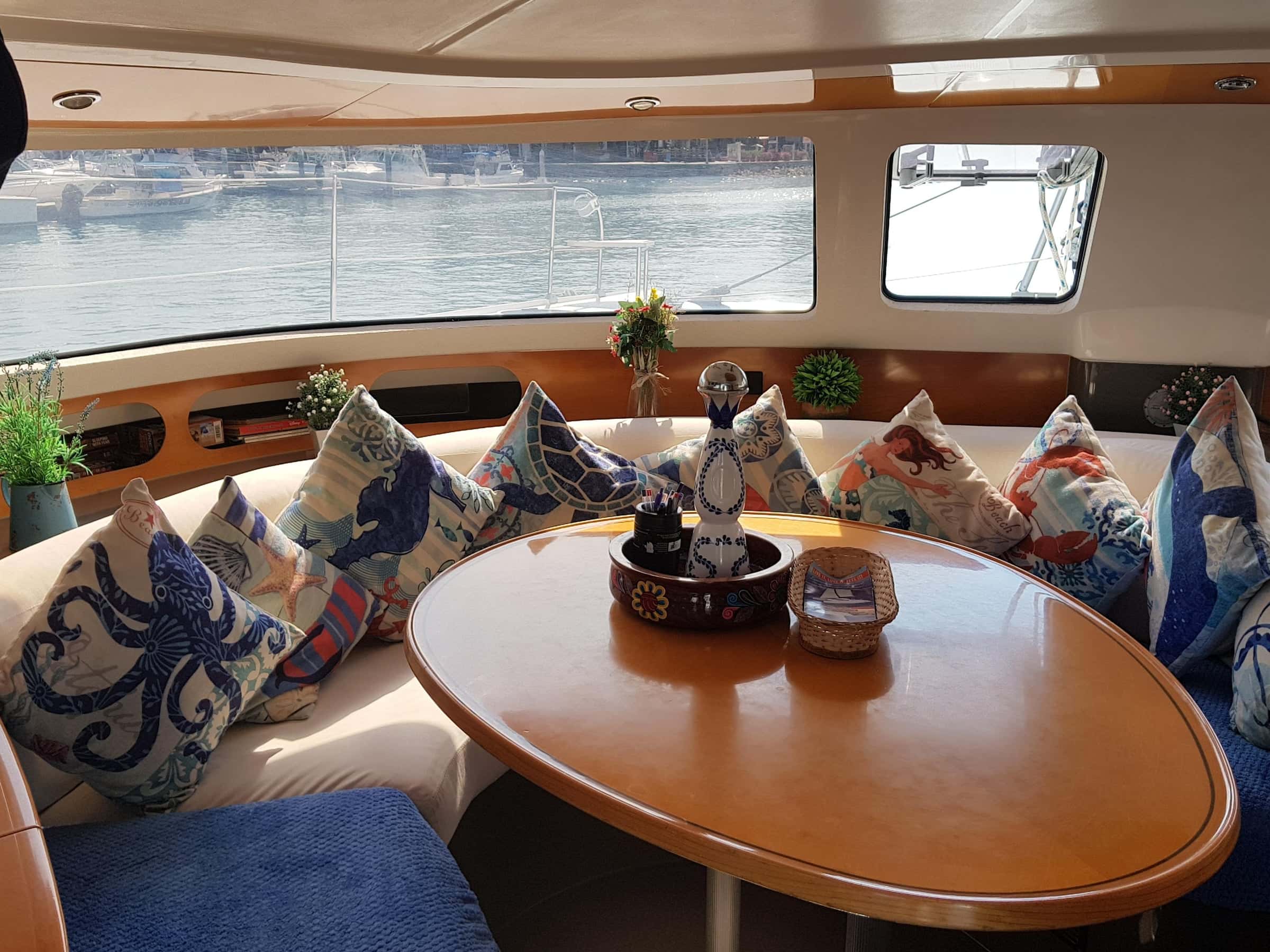 Saloon dinette with an oval wooden table, sea-print cushions and a wide window onto the marina
