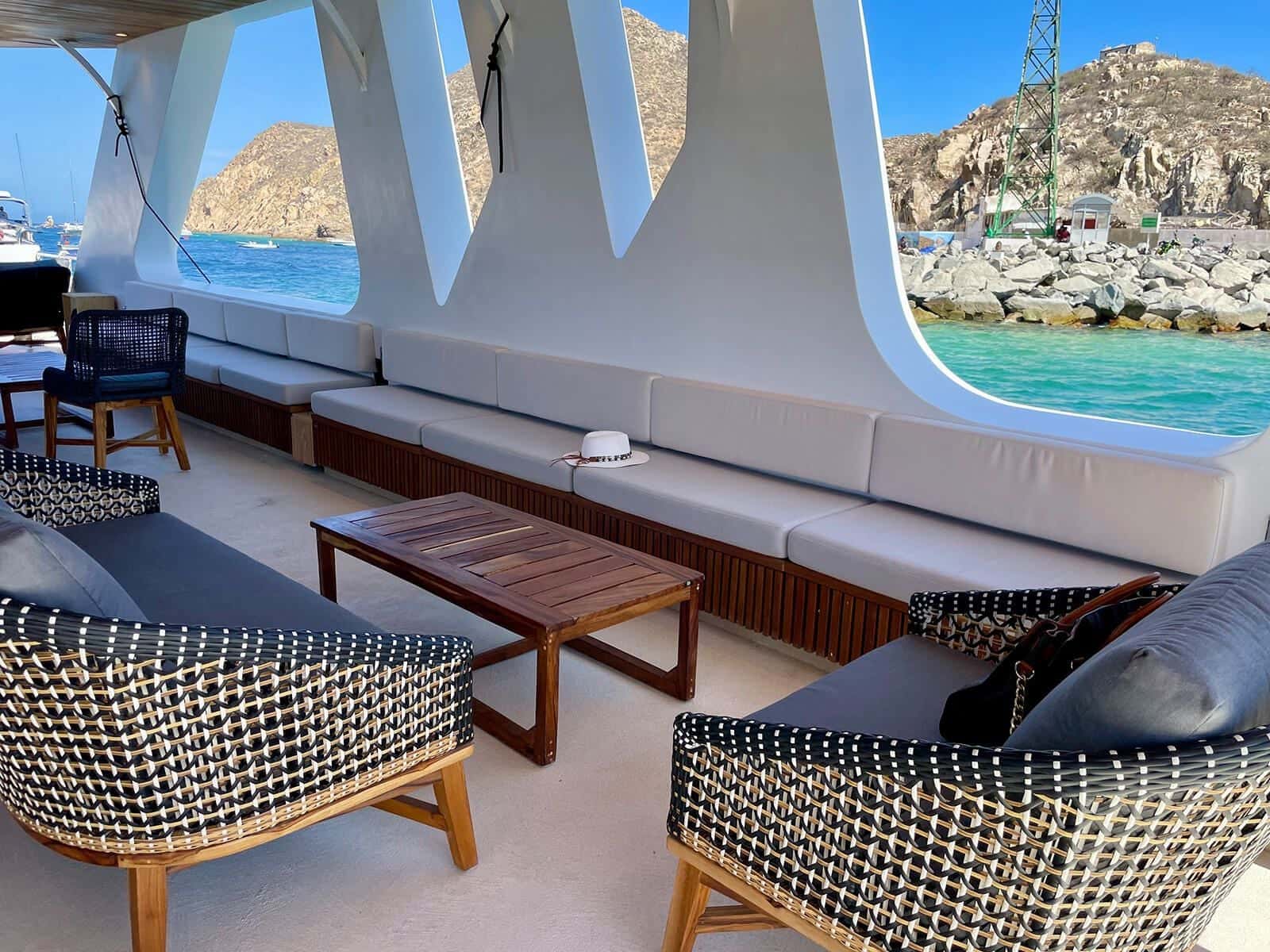 Covered lounge with woven armchairs, a long bench and a low teak table, the bay through the openings