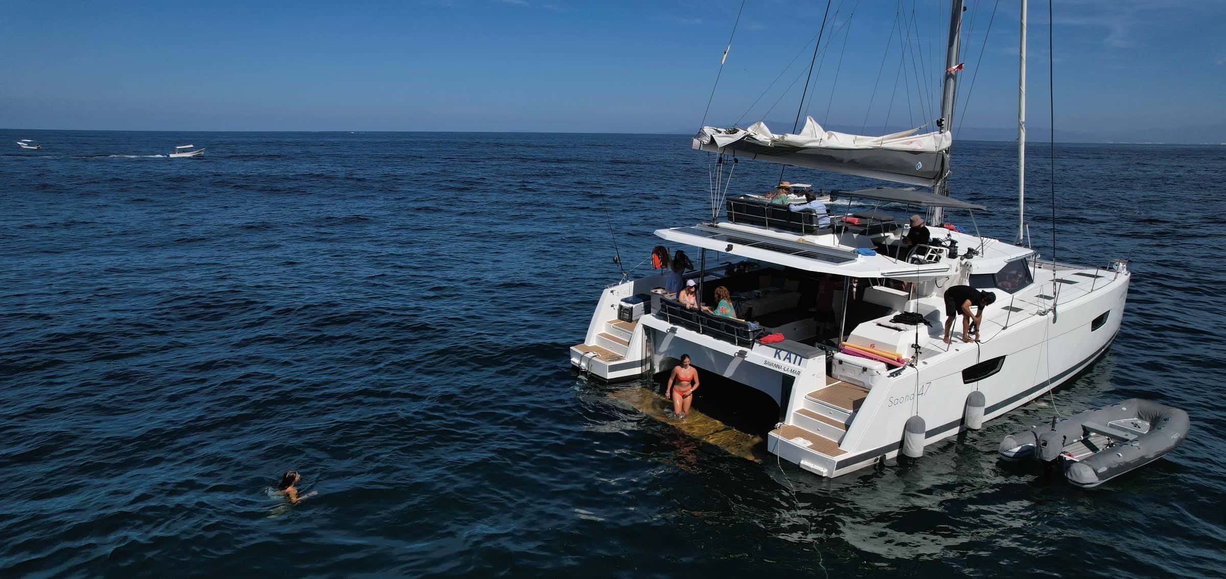 White catamaran seen from astern and above, a swimmer at the stern steps and a grey tender alongside