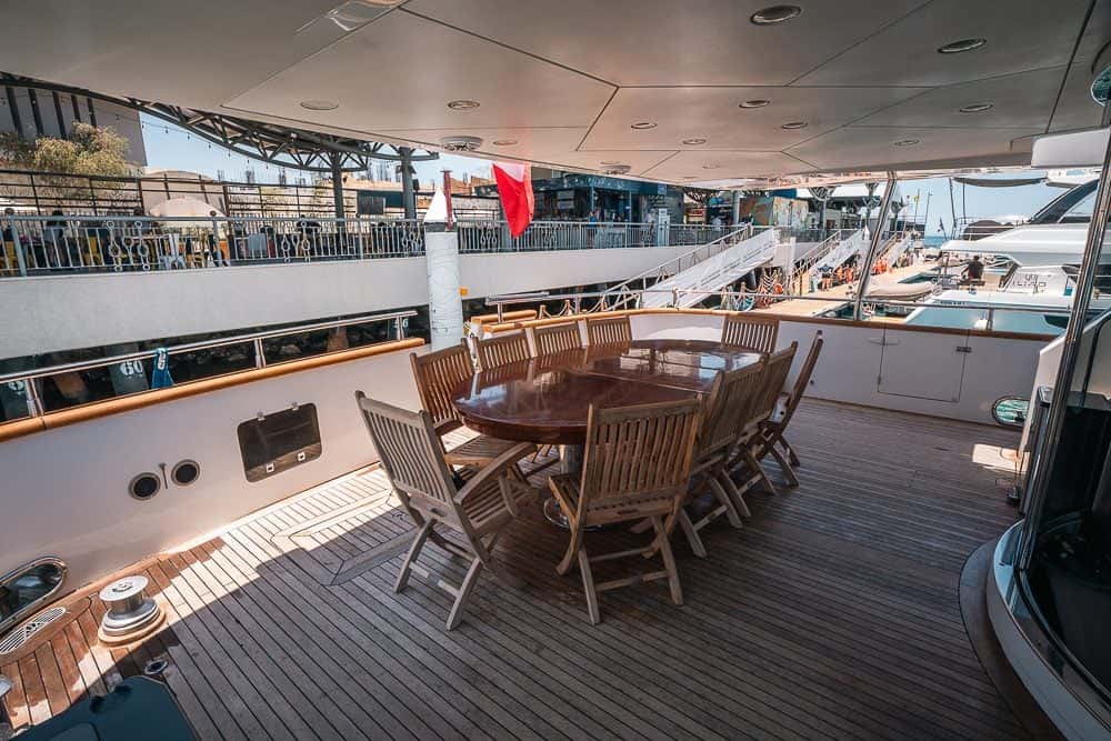 Covered teak aft deck with a long wooden dining table and folding chairs, the marina alongside