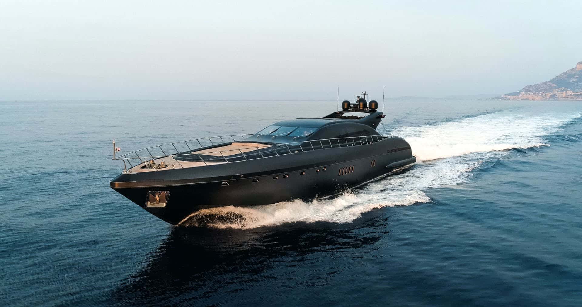 Matte black yacht under way bow-on, a white wake behind and a hillside coast beyond