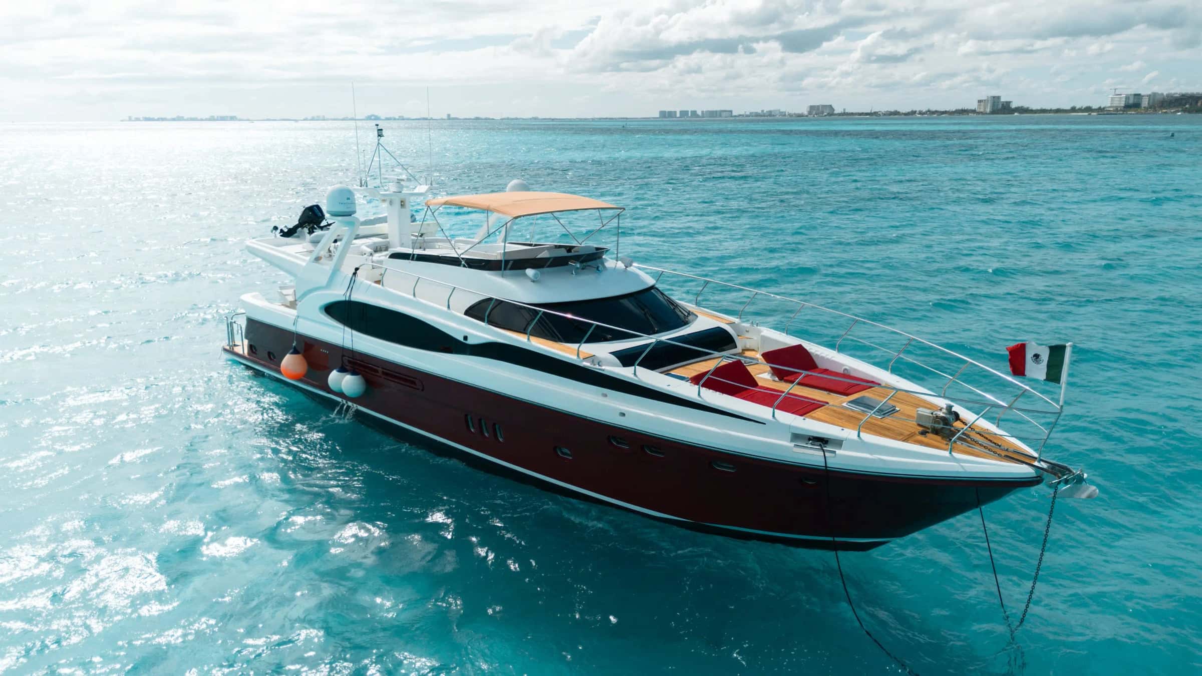 Vive at anchor on turquoise water from the bow quarter, the Mexican flag at the bow and the Cancún shore behind