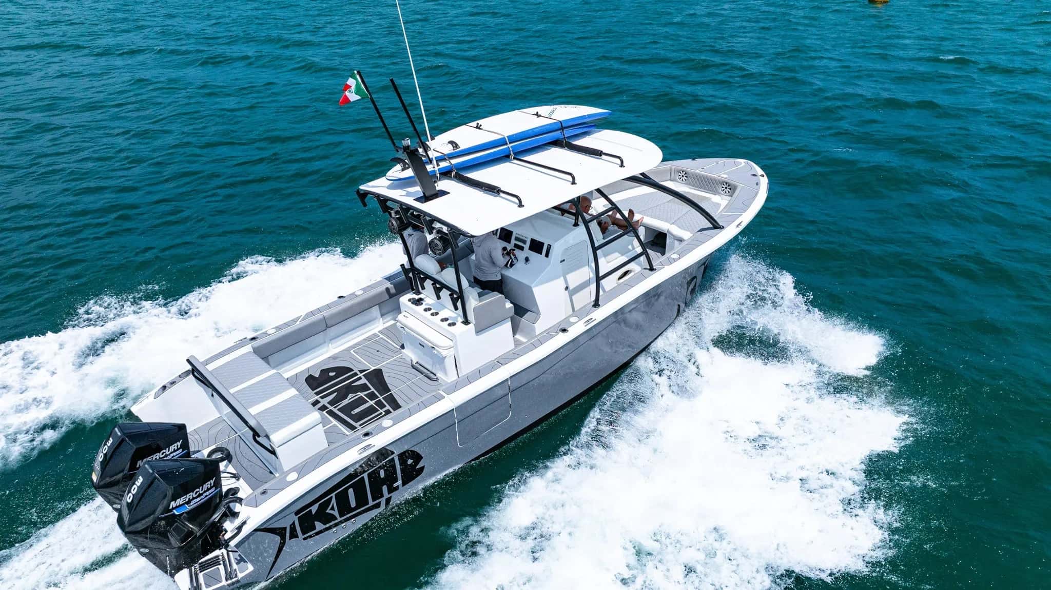Grey sport boat with KOA on the hull and twin black Mercury outboards running at speed, two surfboards strapped to the T-top