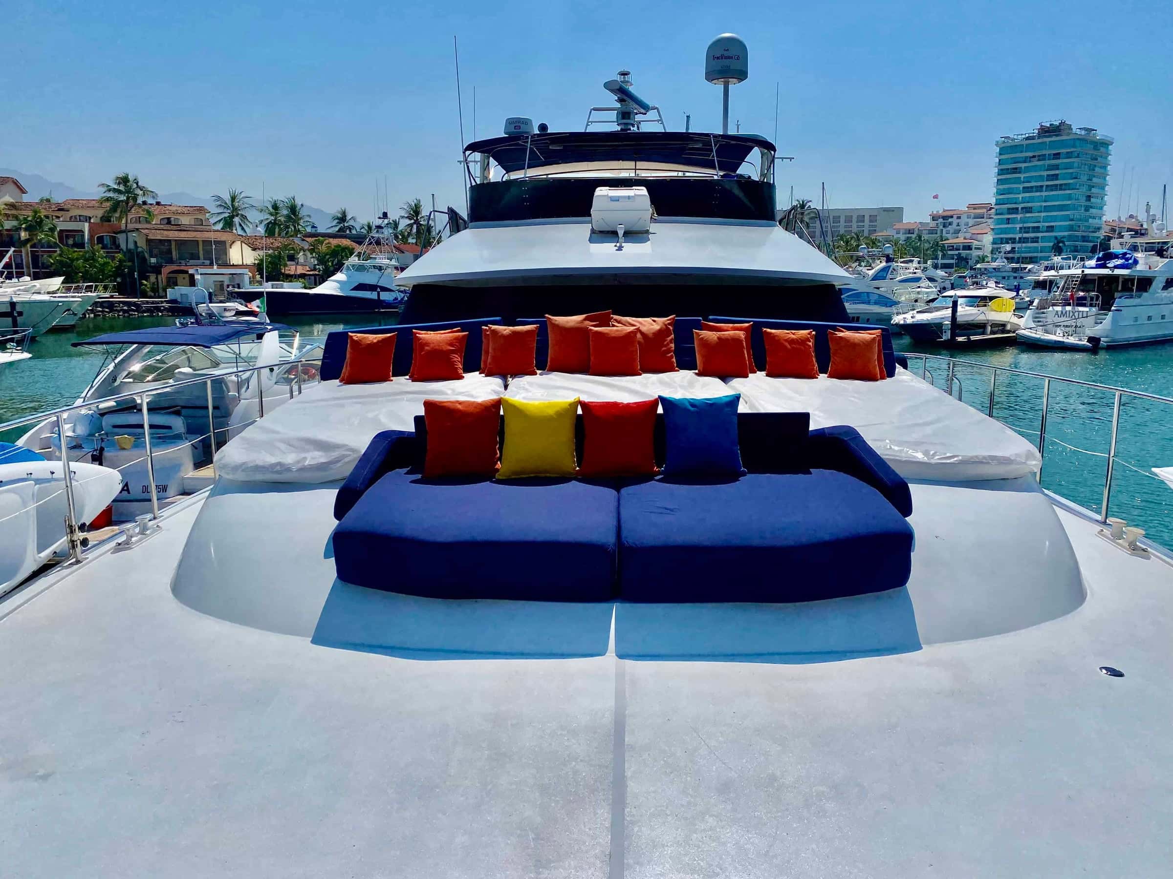 Bow lounge with a blue sofa and red and yellow cushions on a white sun pad, the marina behind