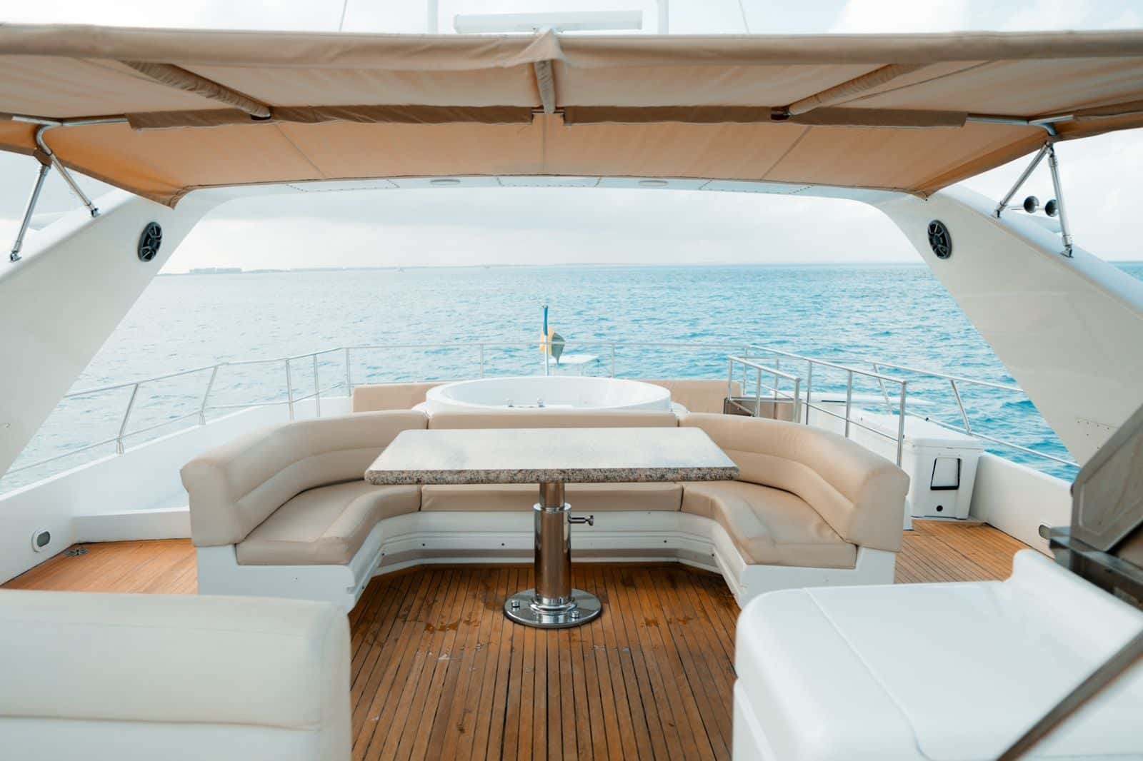 Flybridge lounge with a cream U-shaped sofa and table under a canvas top, the round Jacuzzi beyond and the sea all around