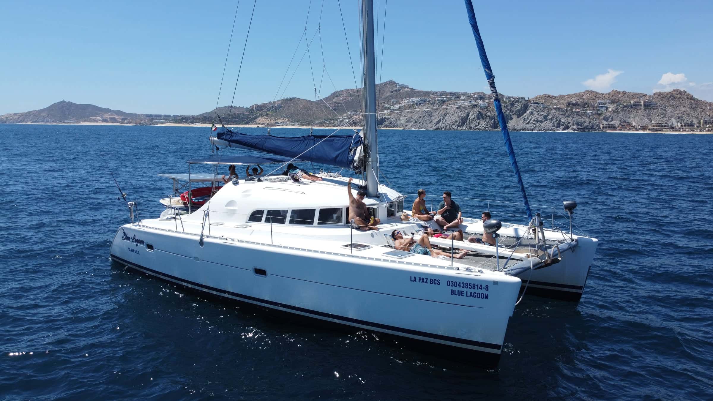 The catamaran at anchor in profile, her name on the hull, the Cabo coast and hills behind