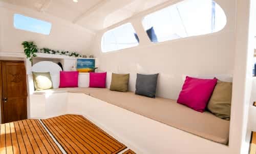 The saloon: a long cushioned bench with bright pillows under the cabin windows, teak steps in the foreground