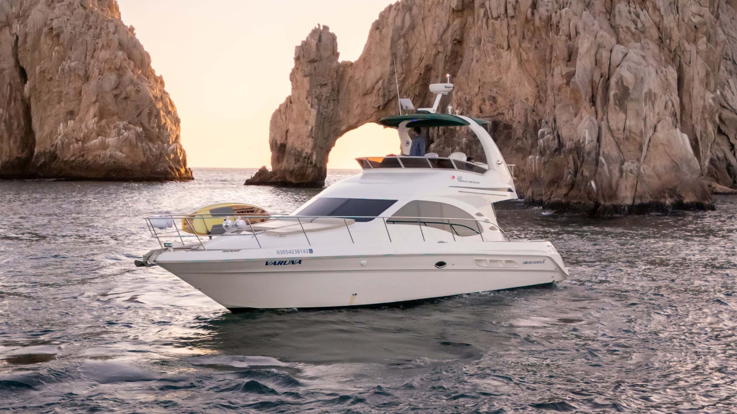 Vaduna, a white Sea Ray flybridge yacht with a green canopy, at rest in front of the Arch at Land's End at sunset