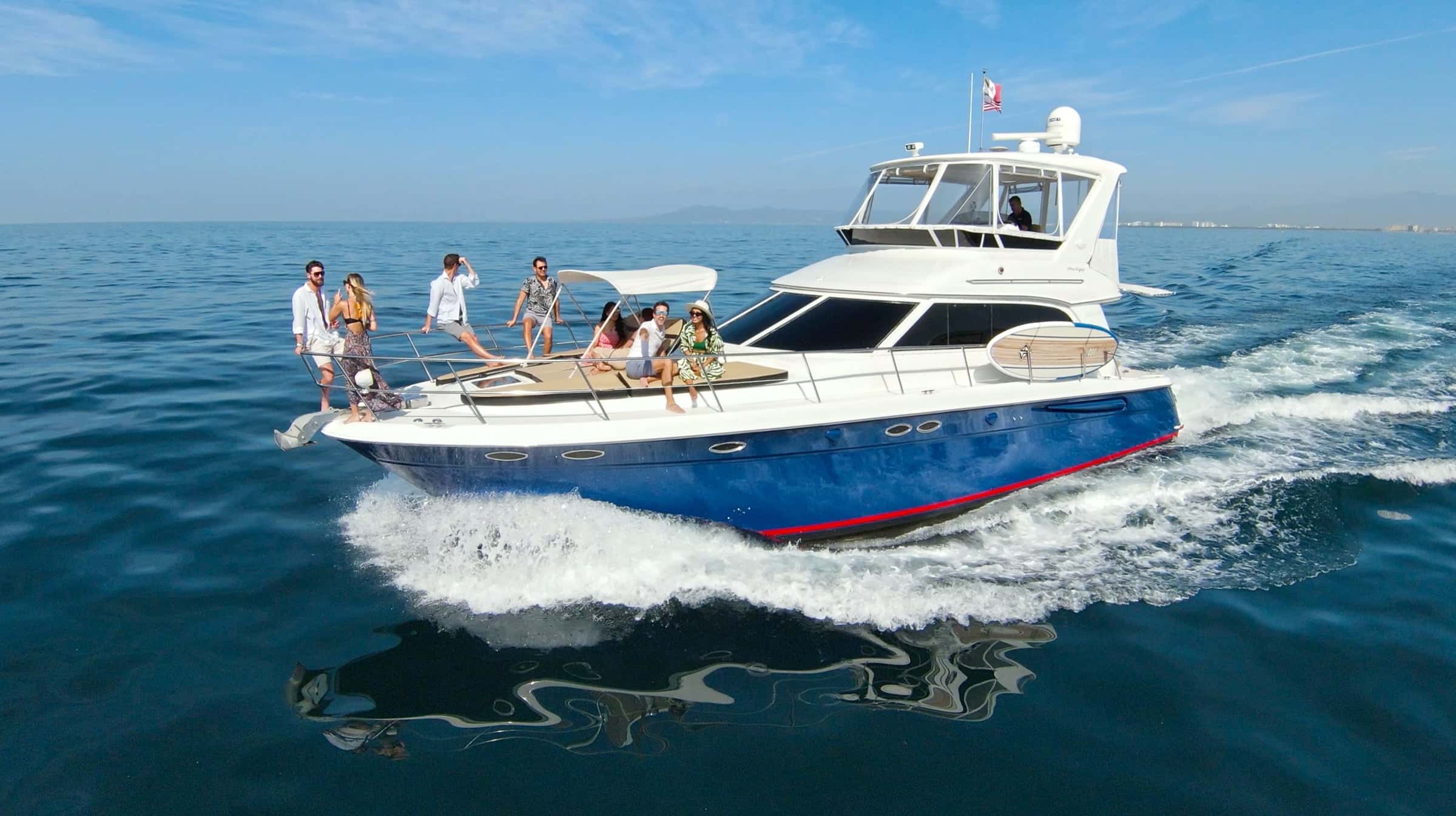 Navy-hulled flybridge motor yacht underway from above, guests standing and sitting on the bow