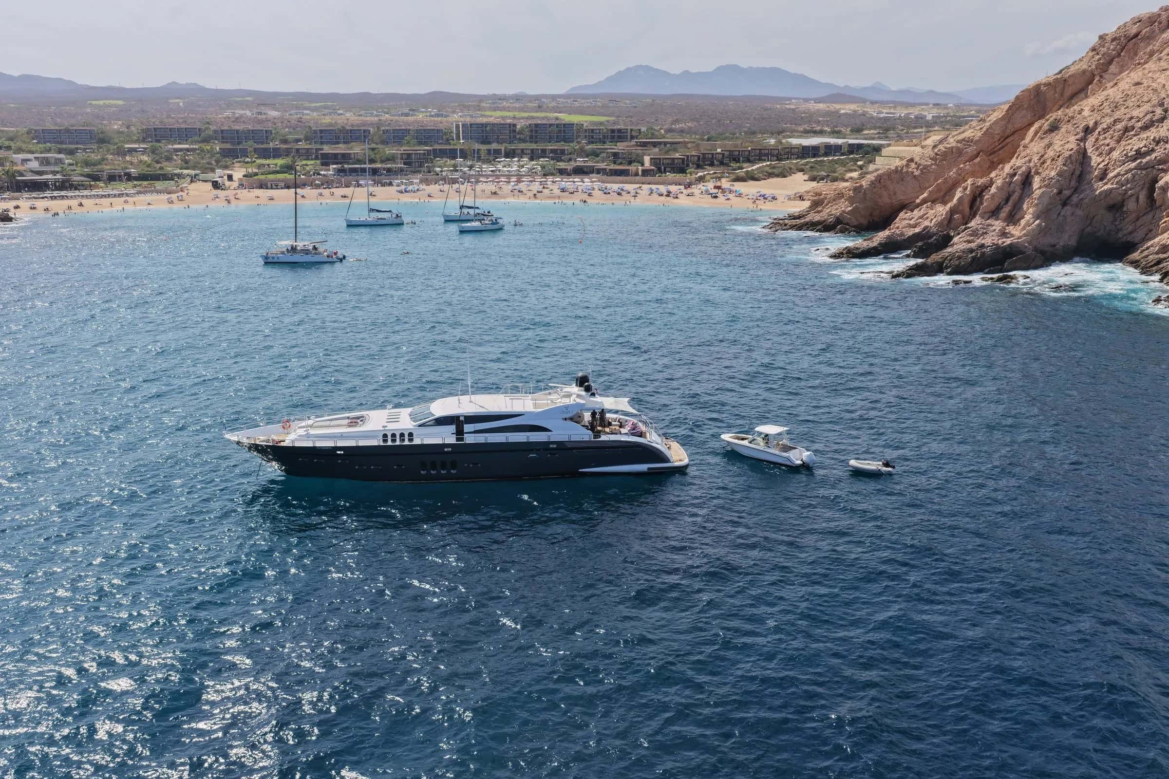 Aerial view of the dark-hulled yacht at anchor in a blue bay, a beach, red rocks and the desert hills behind