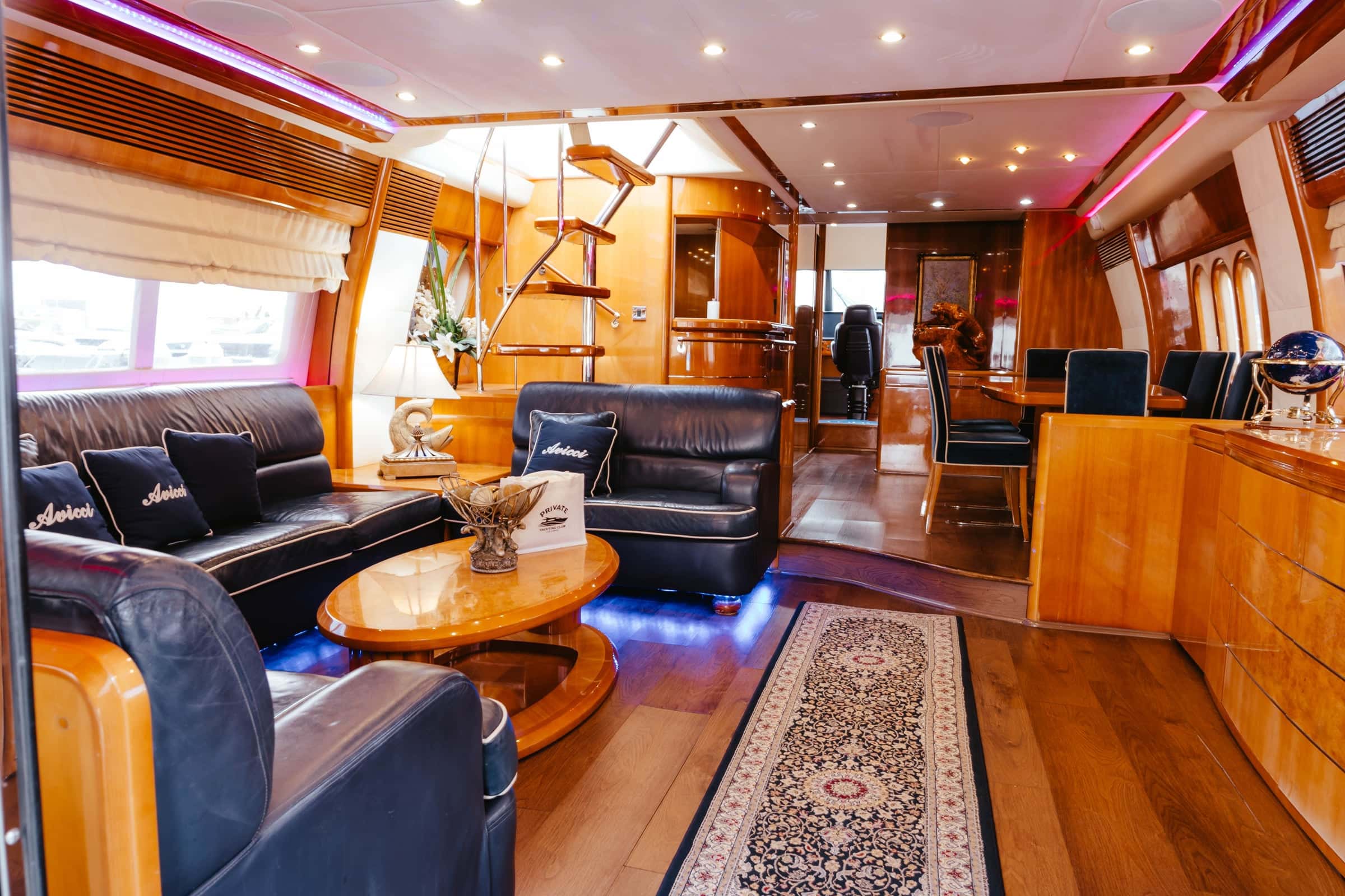 Saloon in polished wood with dark leather sofas, a round coffee table and blue floor lighting, the dining table and galley beyond