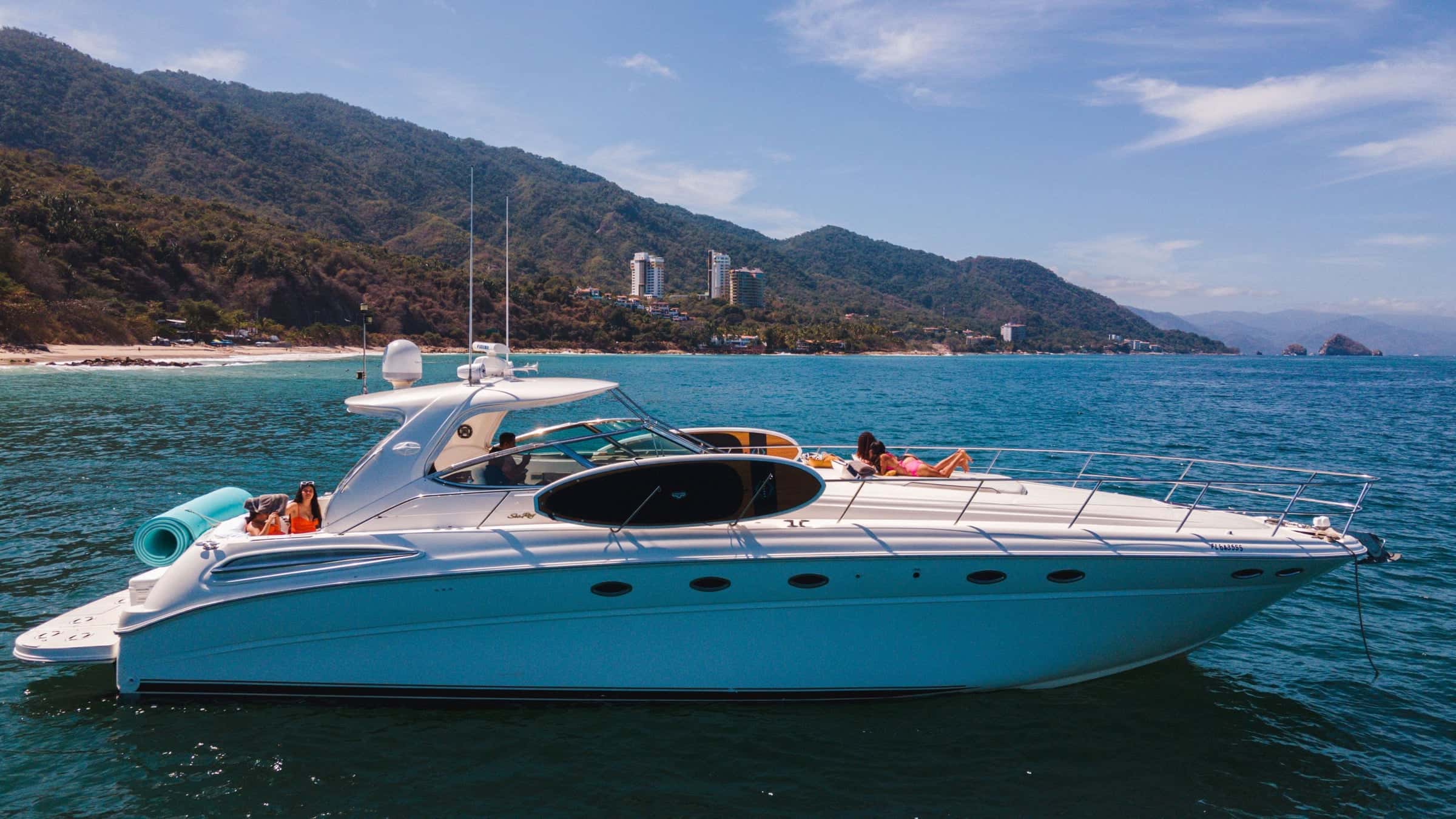 White express motor yacht at anchor from the air, guests on the bow sun pads, green mountains and a beach behind
