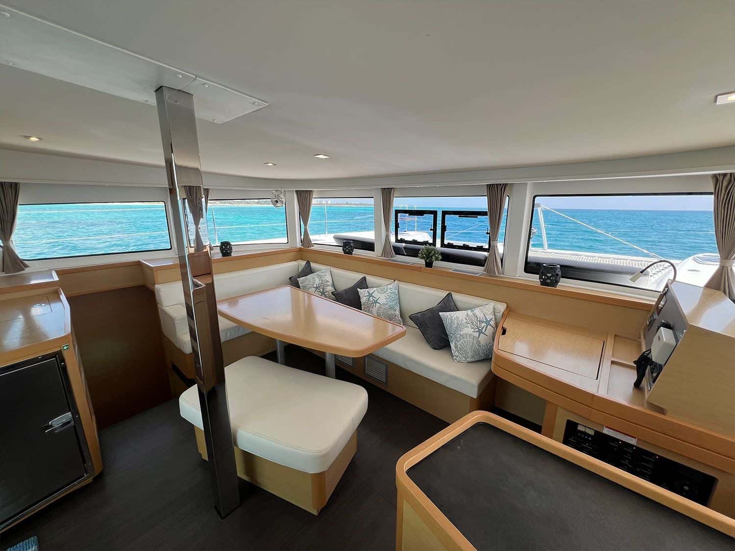 Saloon with a cushioned L-shaped settee around a wooden table, windows all round onto turquoise water