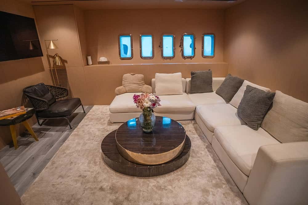 Lounge below deck with an L-shaped cream sofa, grey cushions, a round lacquered coffee table and a row of portholes