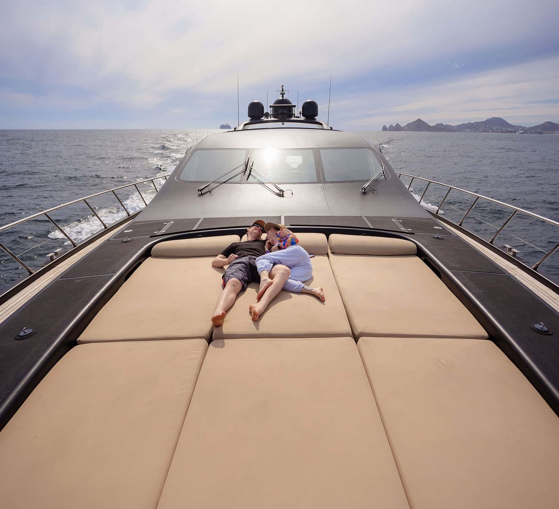 A couple lying on the wide sun pads of a dark yacht's foredeck, the open sea and the Land's End rocks ahead
