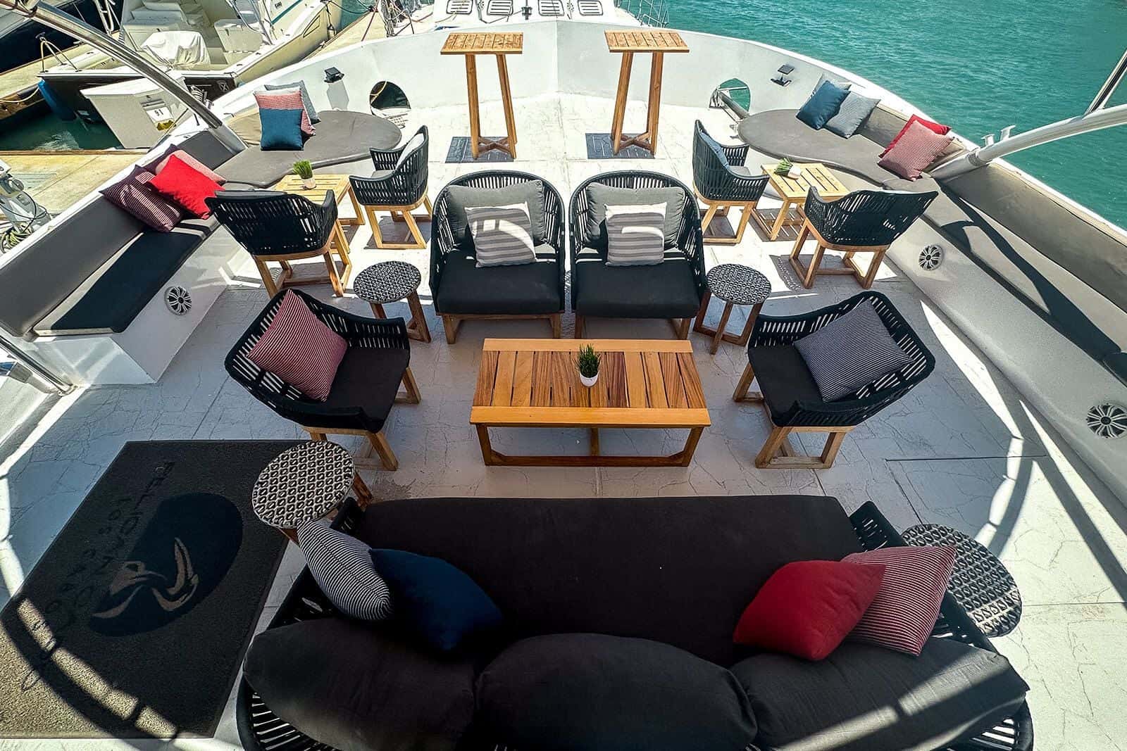 Main deck lounge with woven armchairs, a low teak table and a long sofa with red and blue cushions