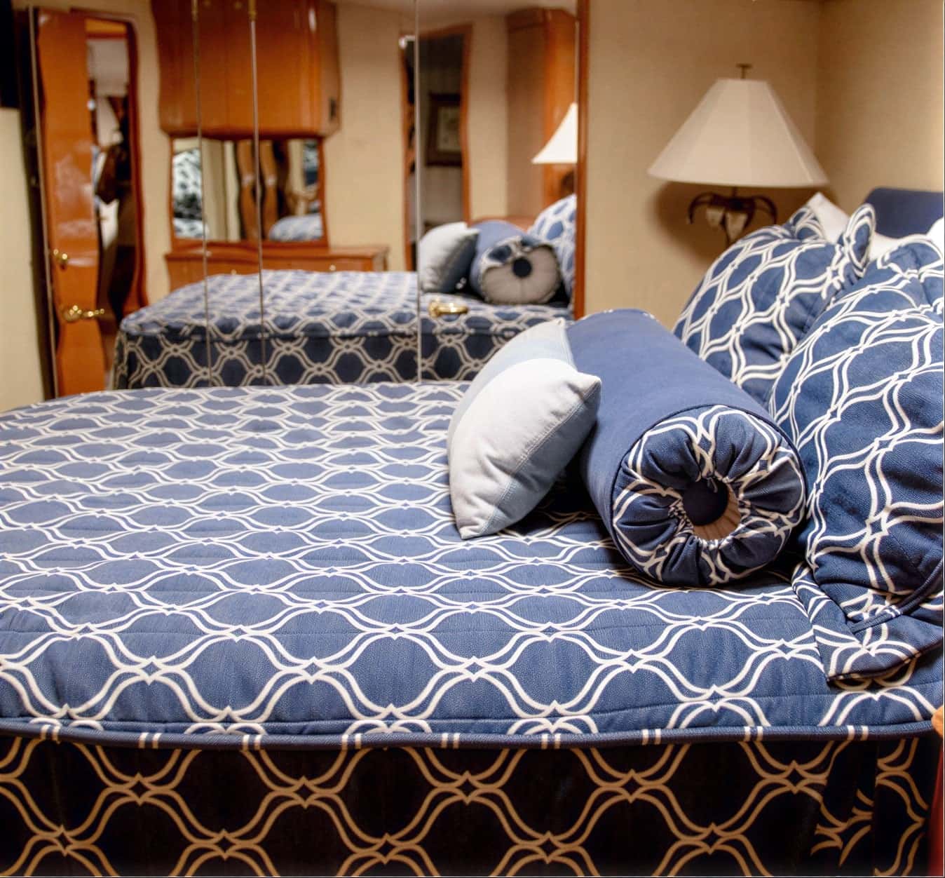 Cabin bed under a blue and white patterned cover with a bolster and cushions, a mirrored wall and a table lamp beside it