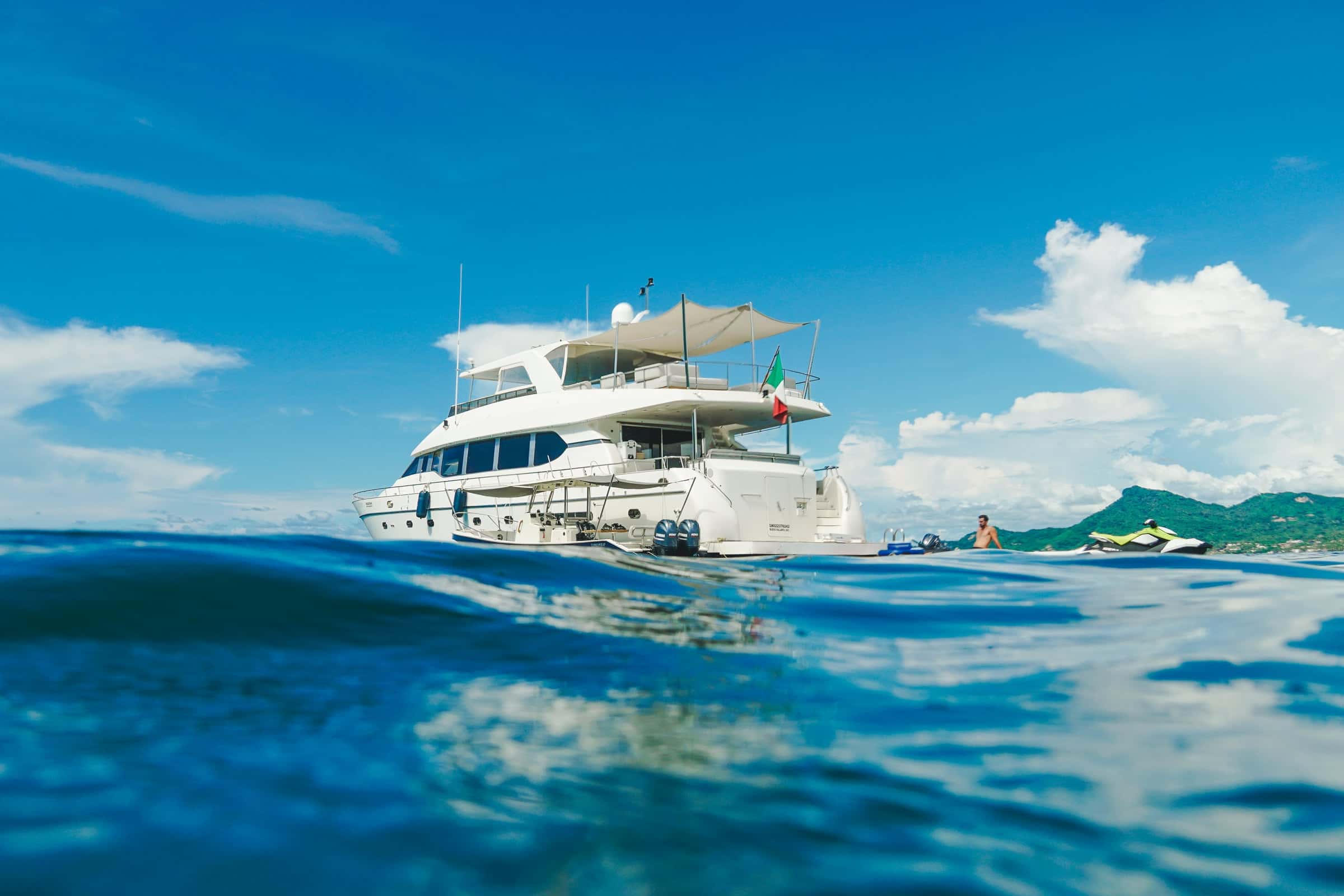 Motor yacht at anchor seen from water level, a jet ski beside her and green hills behind
